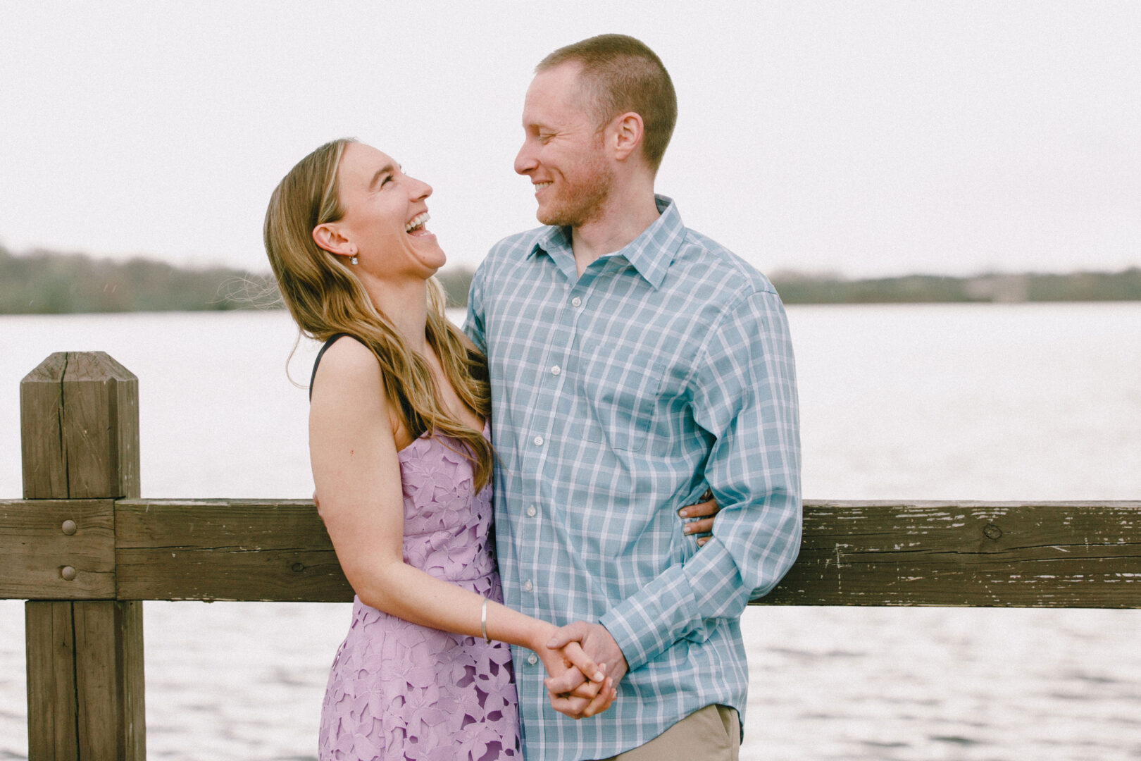 A couple stands by a wooden railing near a body of water, smiling and holding hands.