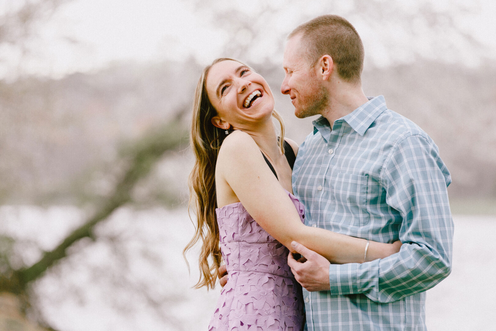 A couple stands outdoors, smiling and embracing, with a blurred natural background.