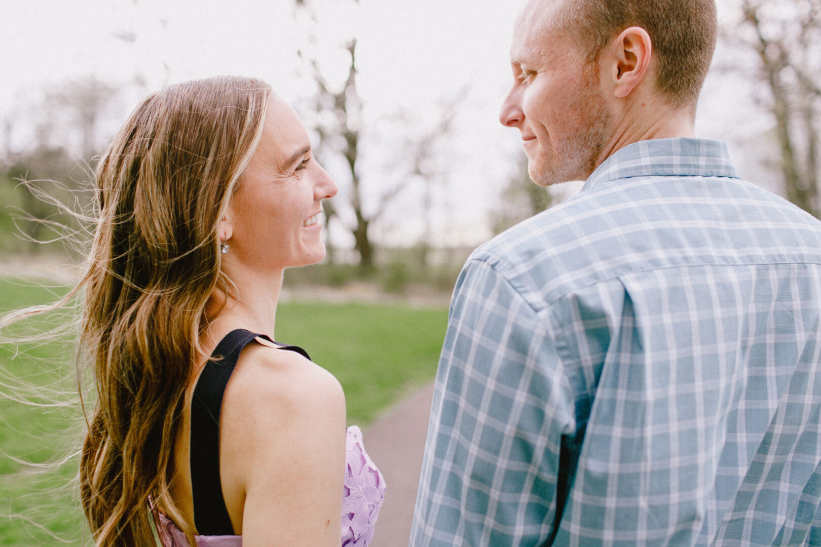 A woman and a man stand outdoors on a path, smiling at each other. Trees and grass are visible in the background.