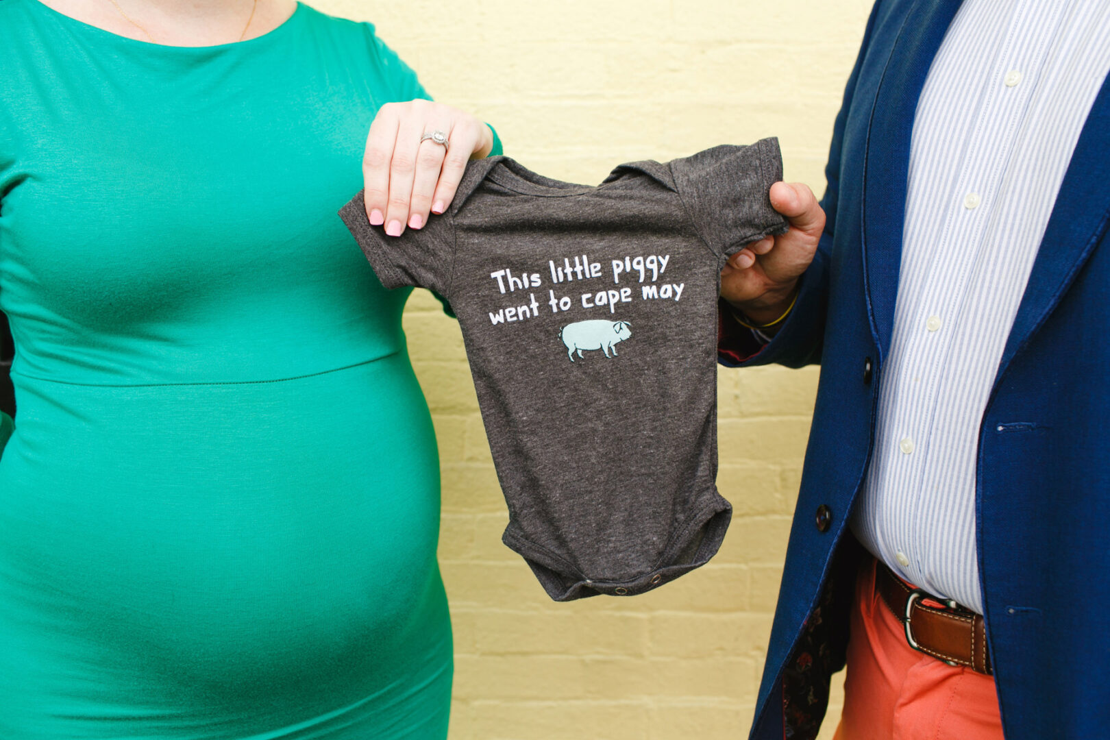 A person in a green dress holds a baby onesie reading "This little piggy went to Cape May" during their Cape May maternity session, standing beside another person in a blue jacket and orange pants.