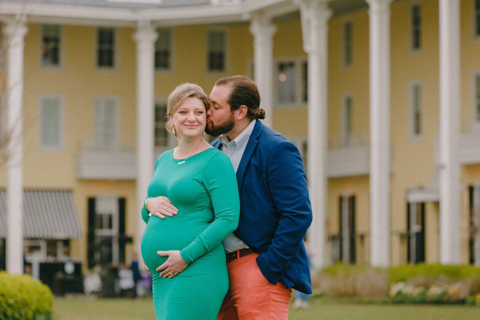 A man gently kisses a pregnant woman's head during a Cape May Maternity Session, standing in front of a yellow building with white pillars.