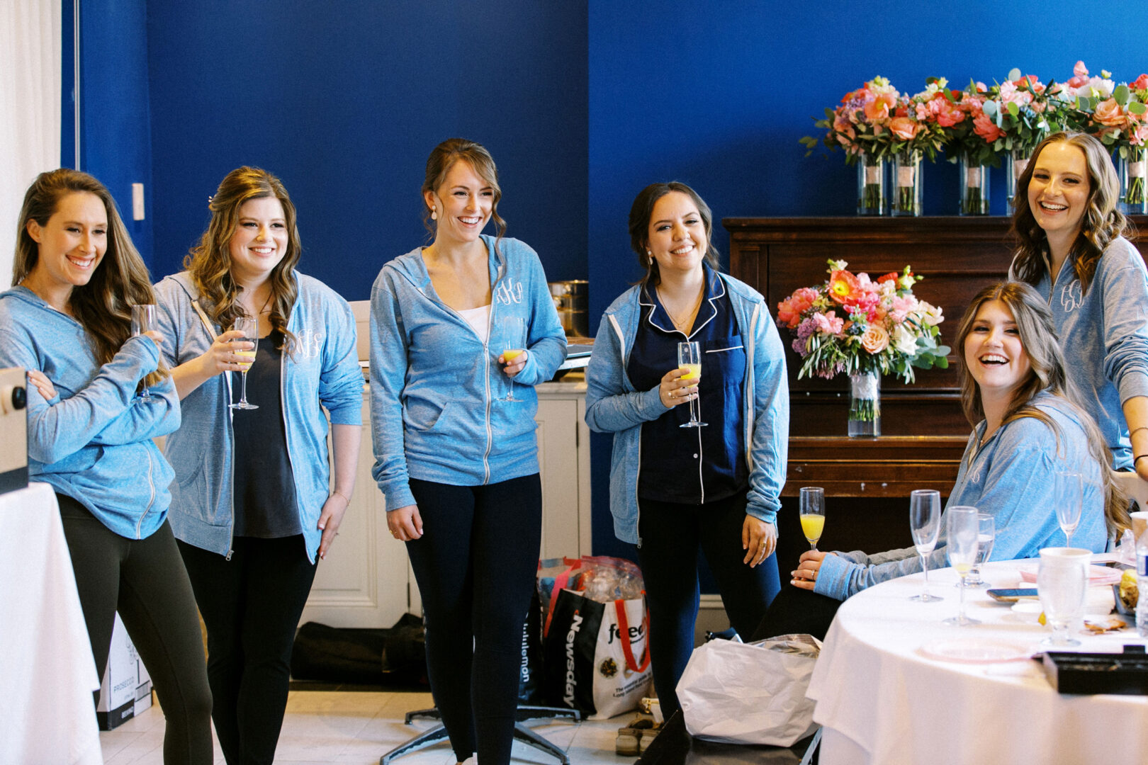 Six women in casual attire, holding champagne glasses, are smiling and talking in a room with blue walls and a table with flowers and plates.