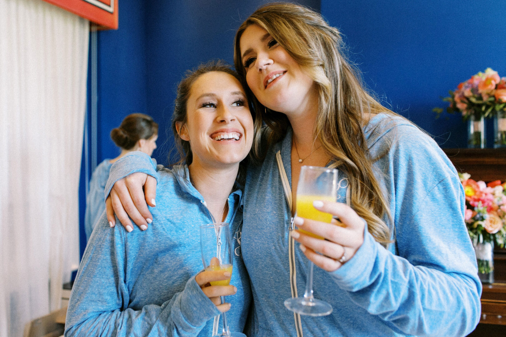 Two women wearing blue tops smile and hold champagne glasses with orange liquid, standing in a room with blue walls and floral arrangements.