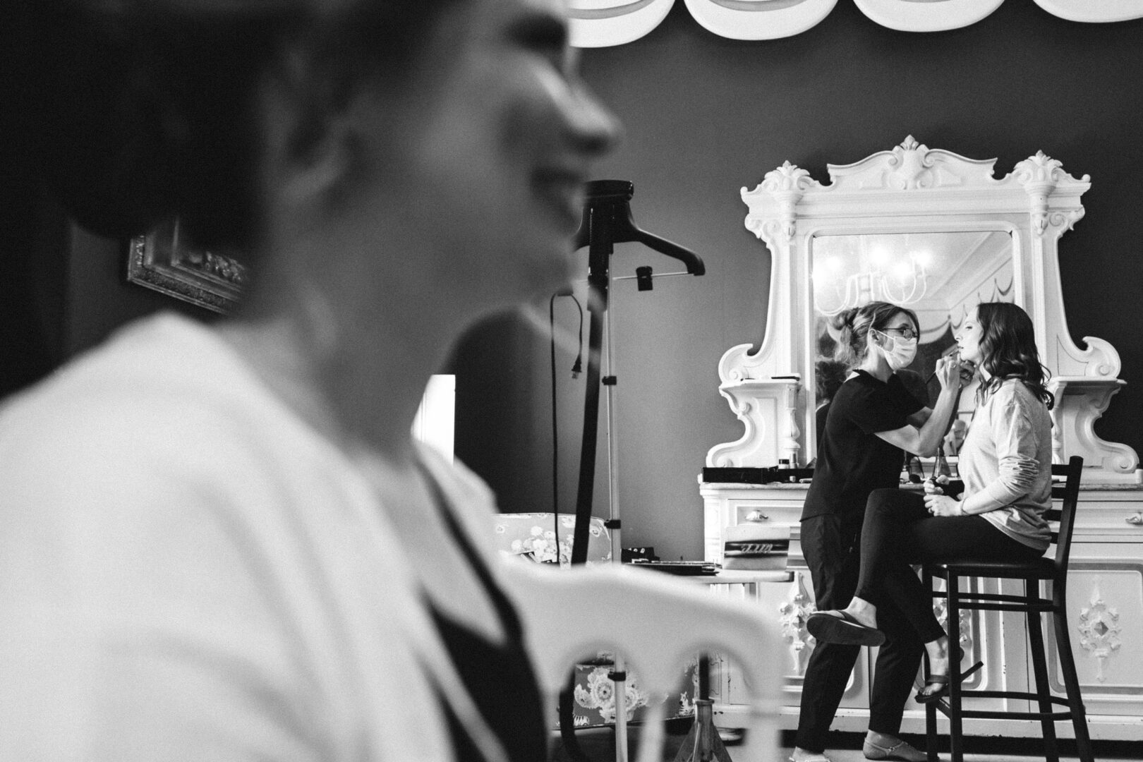 A makeup artist applies makeup to a seated woman in front of a large mirror, while another woman is blurred in the foreground. Black and white image.