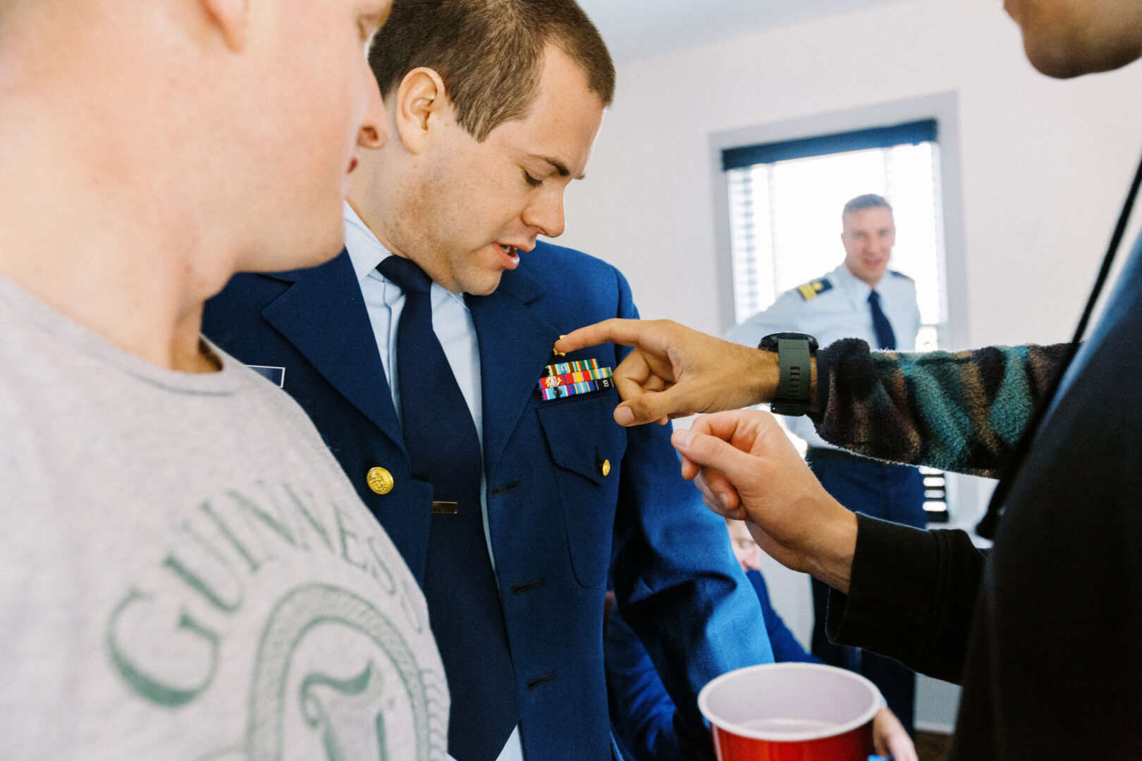 A man in a blue military uniform with ribbons is being shown by others. Another man in the background also wears a white uniform. People are gathered in a casually dressed room.