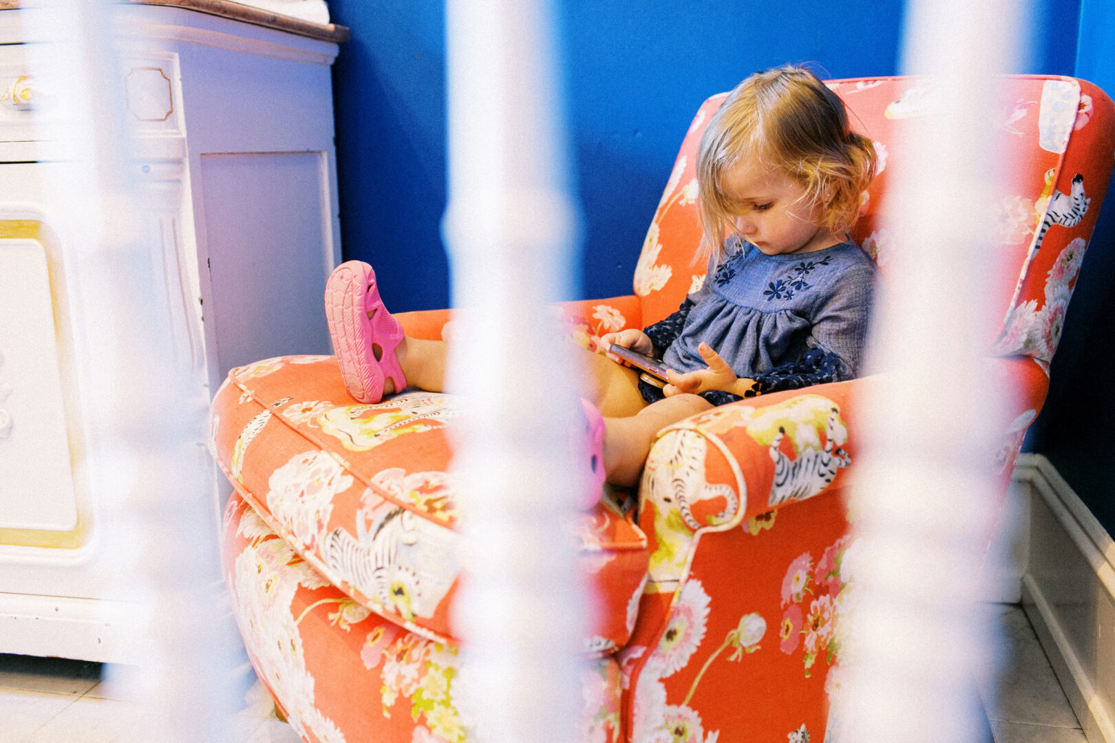 A child sits on a floral armchair, wearing a blue dress and pink shoes, focused on a tablet in a room with blue walls. White bars are visible in the foreground, reminiscent of the serene backdrop you'd find at a wedding by the beach in Cape May.