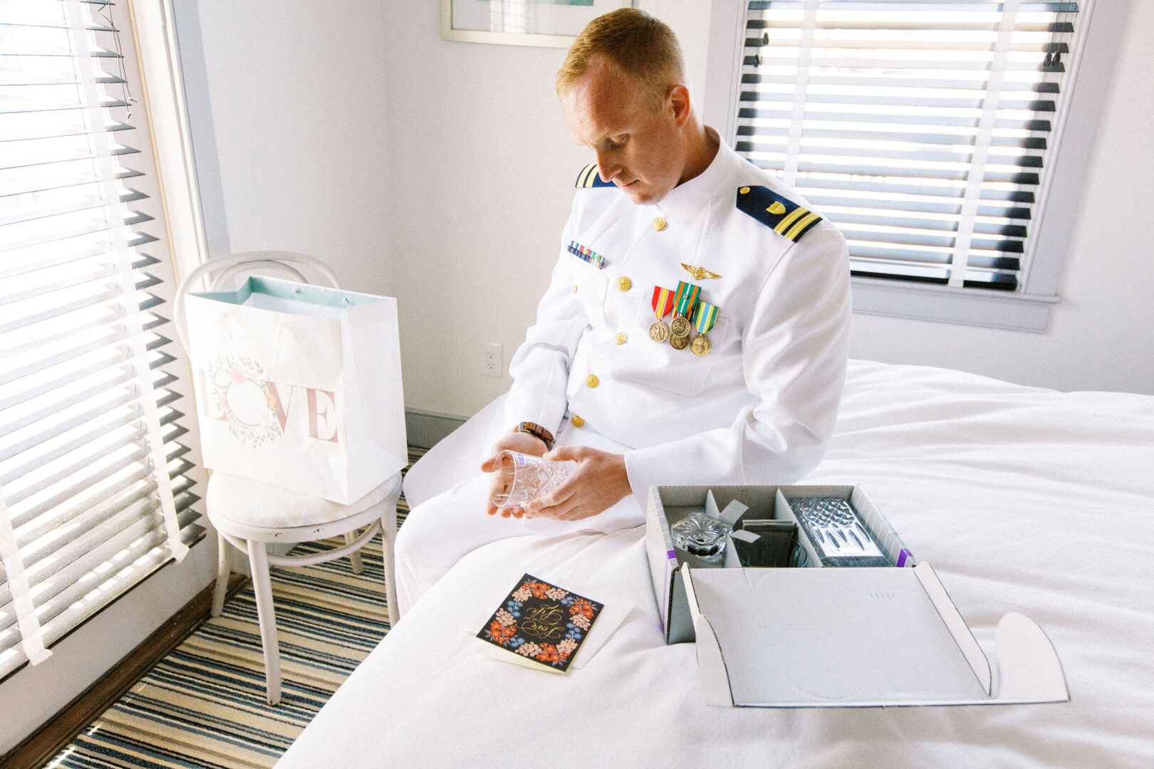A man in a military dress uniform sits on a bed, holding a small box, likely an engagement ring for their upcoming wedding by the beach in Cape May. Nearby, there is a gift bag and a book laying on the bed.