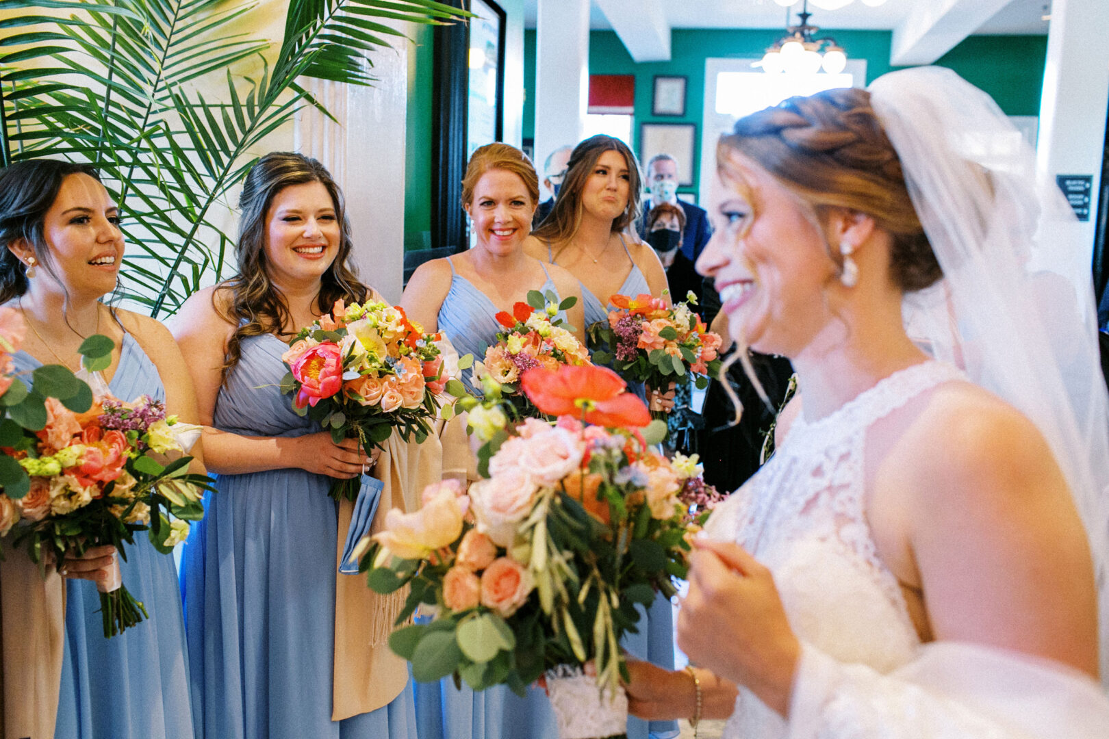 A bride in a white dress and veil stands by the beach in Cape May, holding a bouquet and smiling at five bridesmaids in blue dresses, each clutching colorful bouquets.