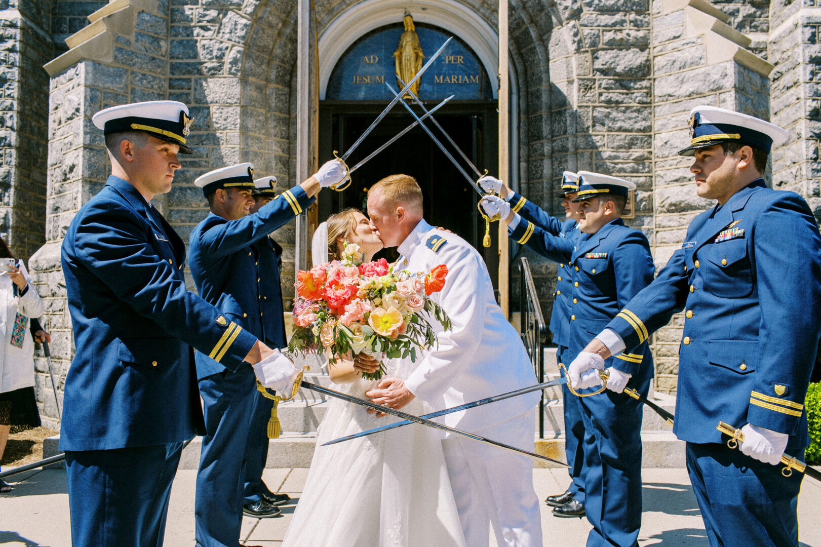 A couple kisses under an arch of swords, formed by uniformed officers outside a stone building, echoing the charm of a wedding by the beach in Cape May. The bride holds a bouquet of colorful flowers.