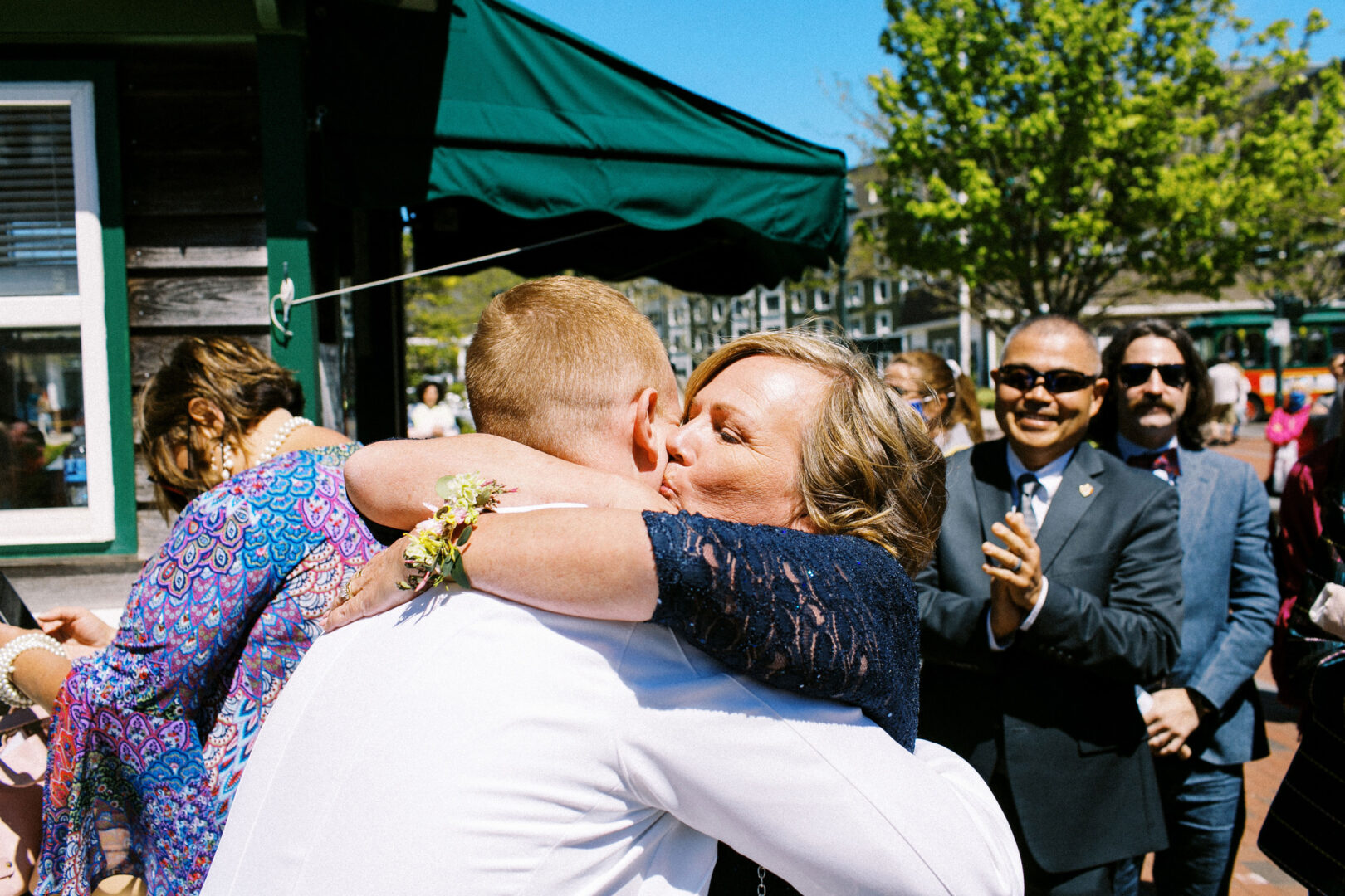 A woman in a dark dress embraces a man in a white shirt outdoors during a beautiful wedding by the beach in Cape May, as people in the background applaud.