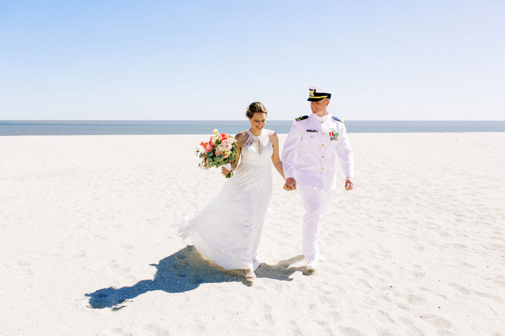 A couple in wedding attire strolls along a sandy beach in Cape May; the bride holds a bouquet, and the groom is dressed in a naval uniform.