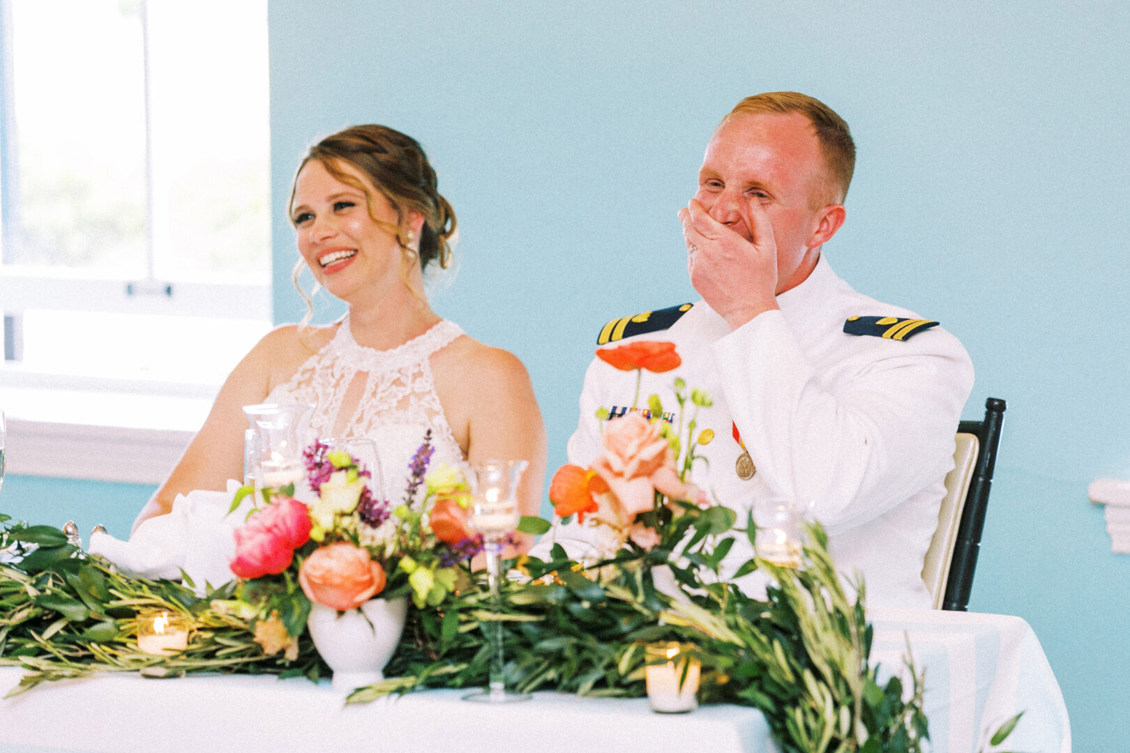A couple sits smiling at a decorated table by the beach in Cape May, with the man in a military uniform and the woman in a white dress. They are surrounded by colorful flowers and greenery, capturing the essence of their coastal wedding.