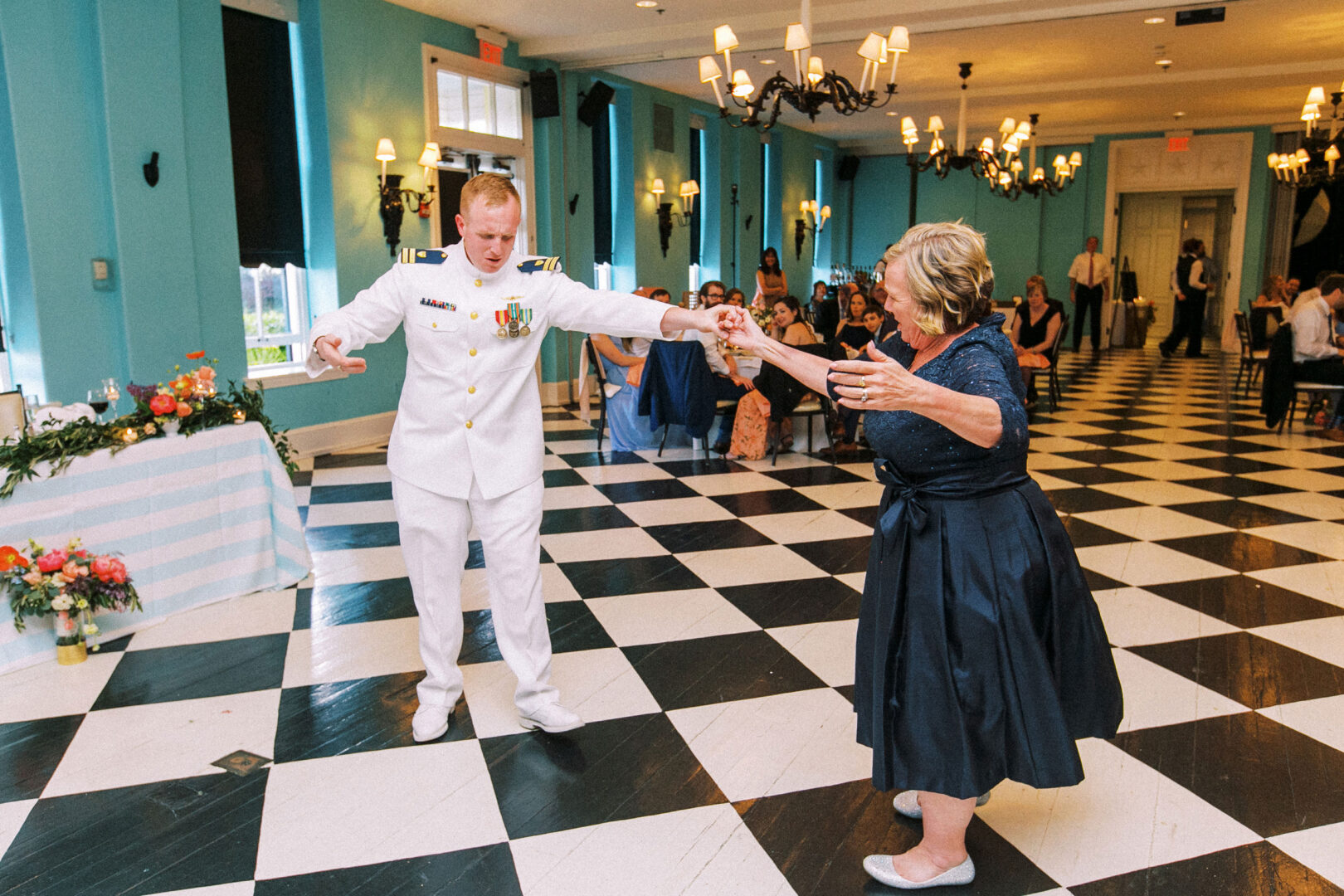 A person in a naval uniform dances with an older woman on a black and white checkered floor in a bright room, reminiscent of a wedding by the beach in Cape May. Other guests are seated in the background.