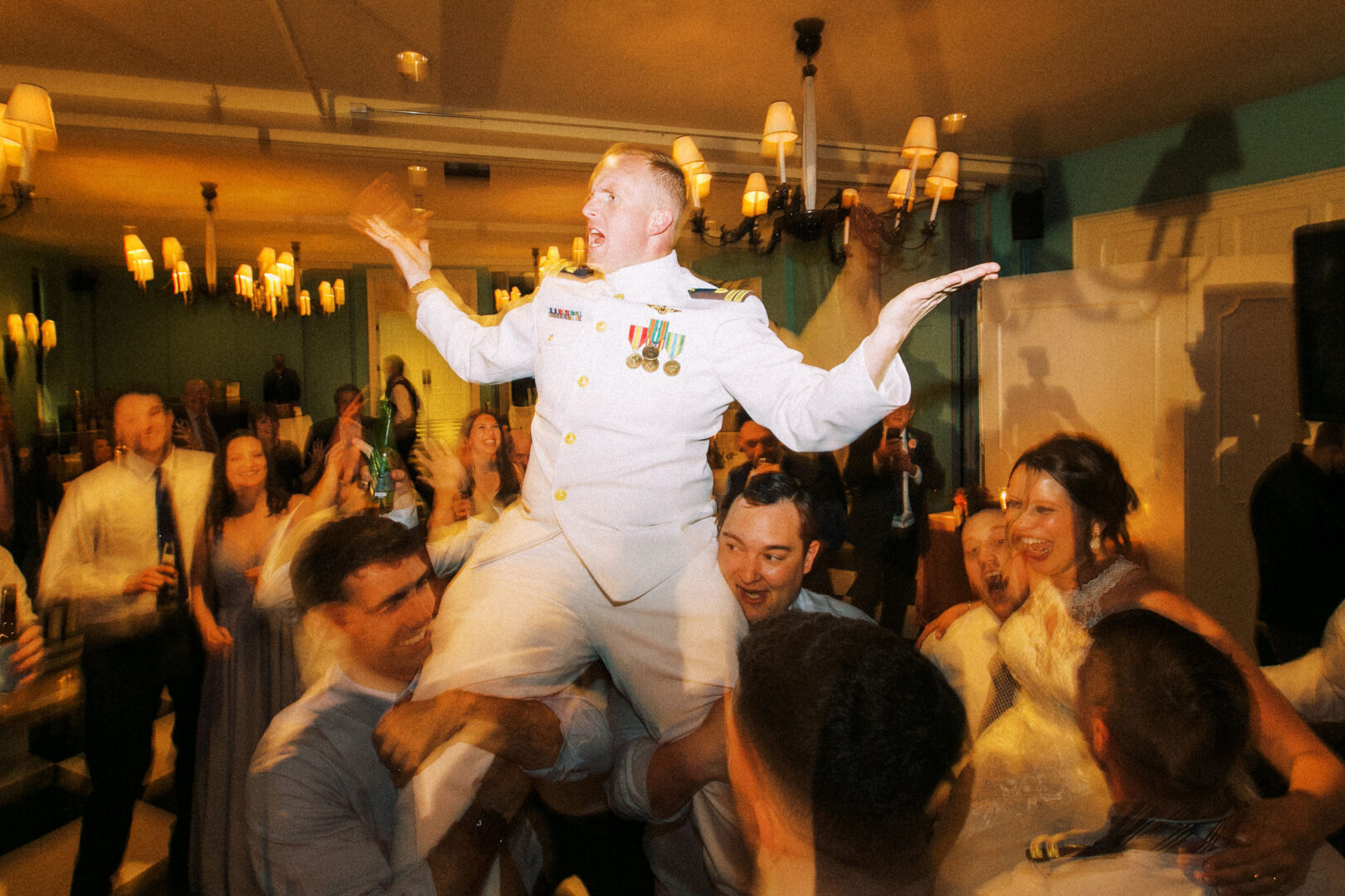 A person in a naval uniform is lifted by a jubilant group at a lively indoor celebration, reminiscent of a festive wedding by the beach in Cape May.