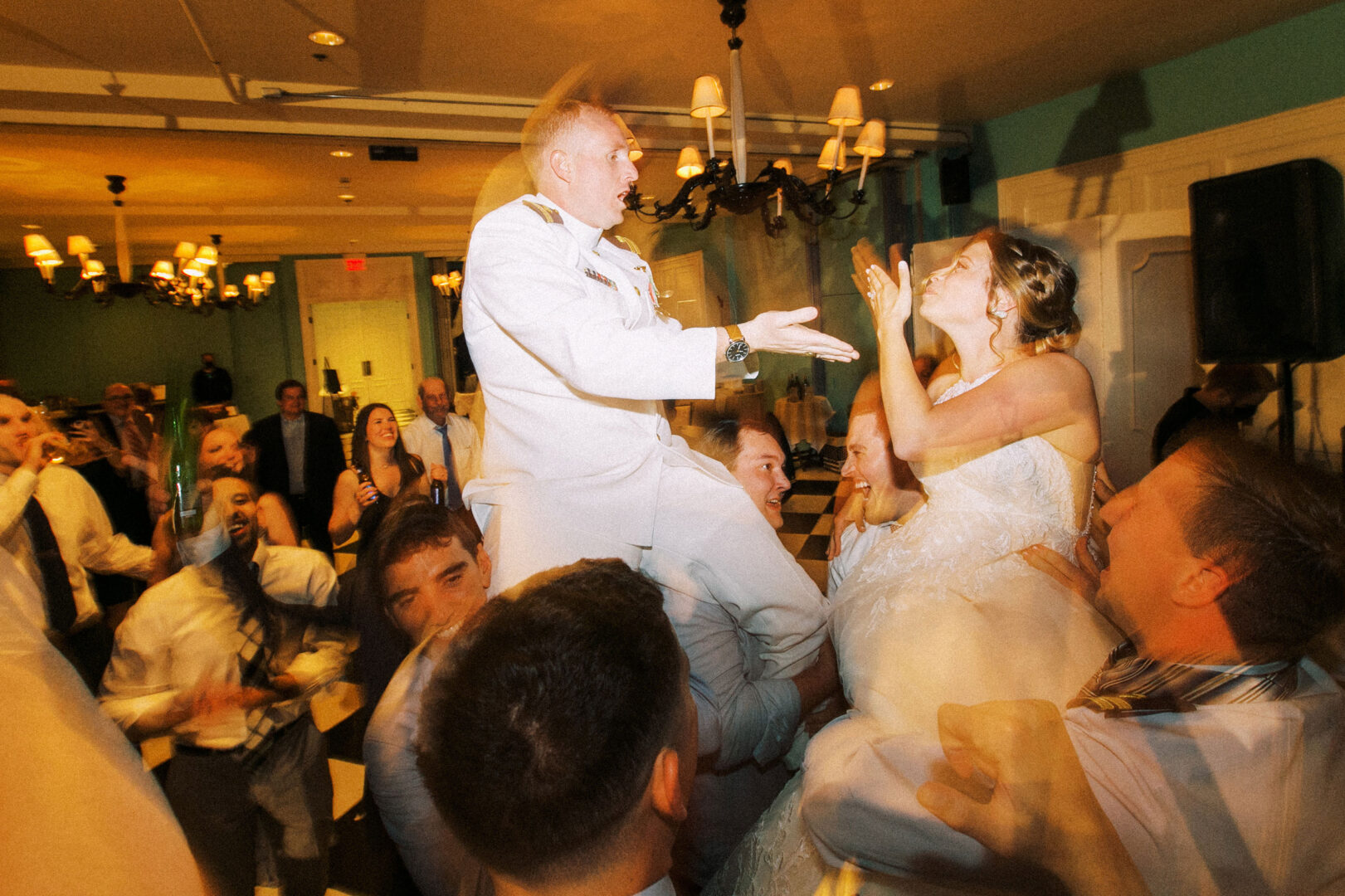 Bride and groom are lifted in chairs by guests during a lively wedding celebration in a warmly lit room, reminiscent of a joyous gathering at a wedding by the beach in Cape May.