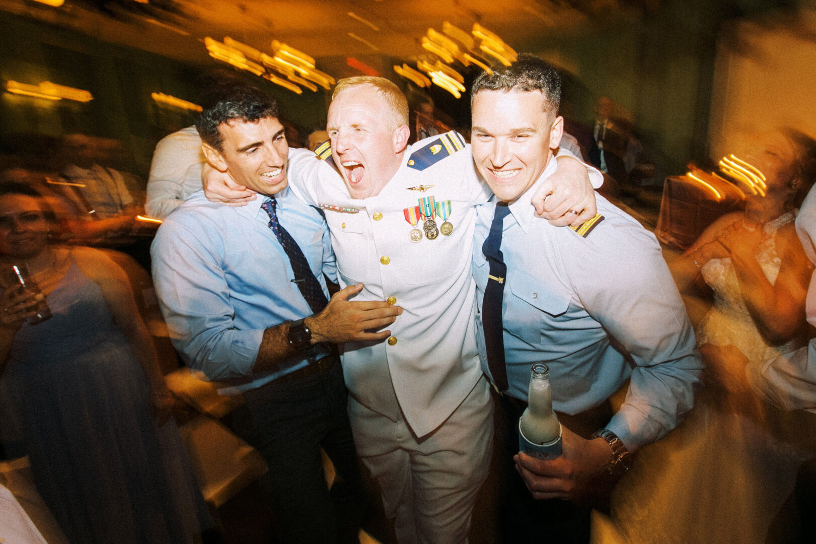Three men in a lively moment at a beach wedding in Cape May; one in a white military uniform, flanked by two men in shirts and ties, celebrating with smiles and casual drinks in hand.