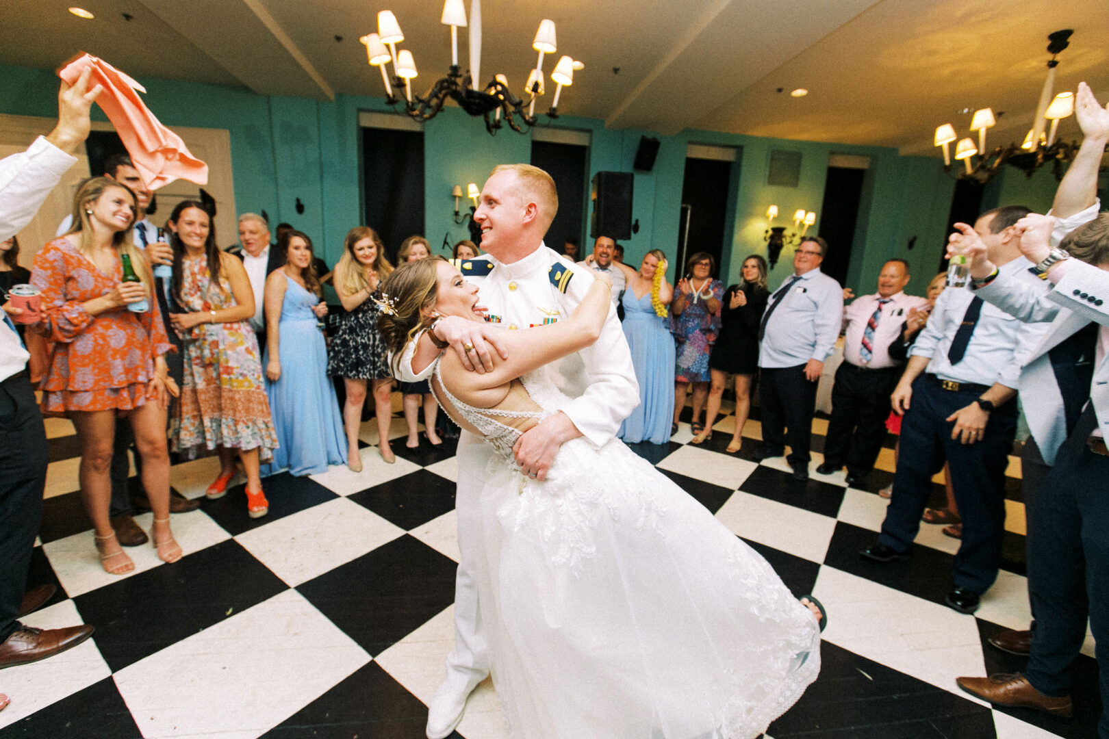 At a wedding by the beach in Cape May, a couple in formal attire dances joyfully on a black-and-white checkered floor, surrounded by clapping and smiling guests.