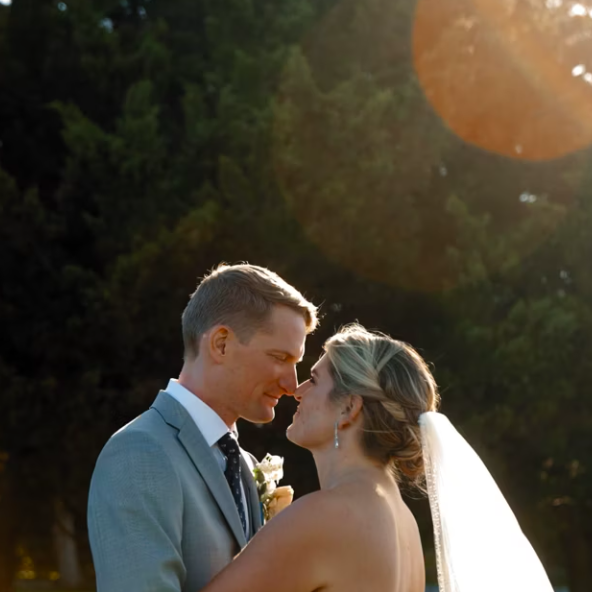 A couple in wedding attire embraces outdoors under sunlight, surrounded by trees.