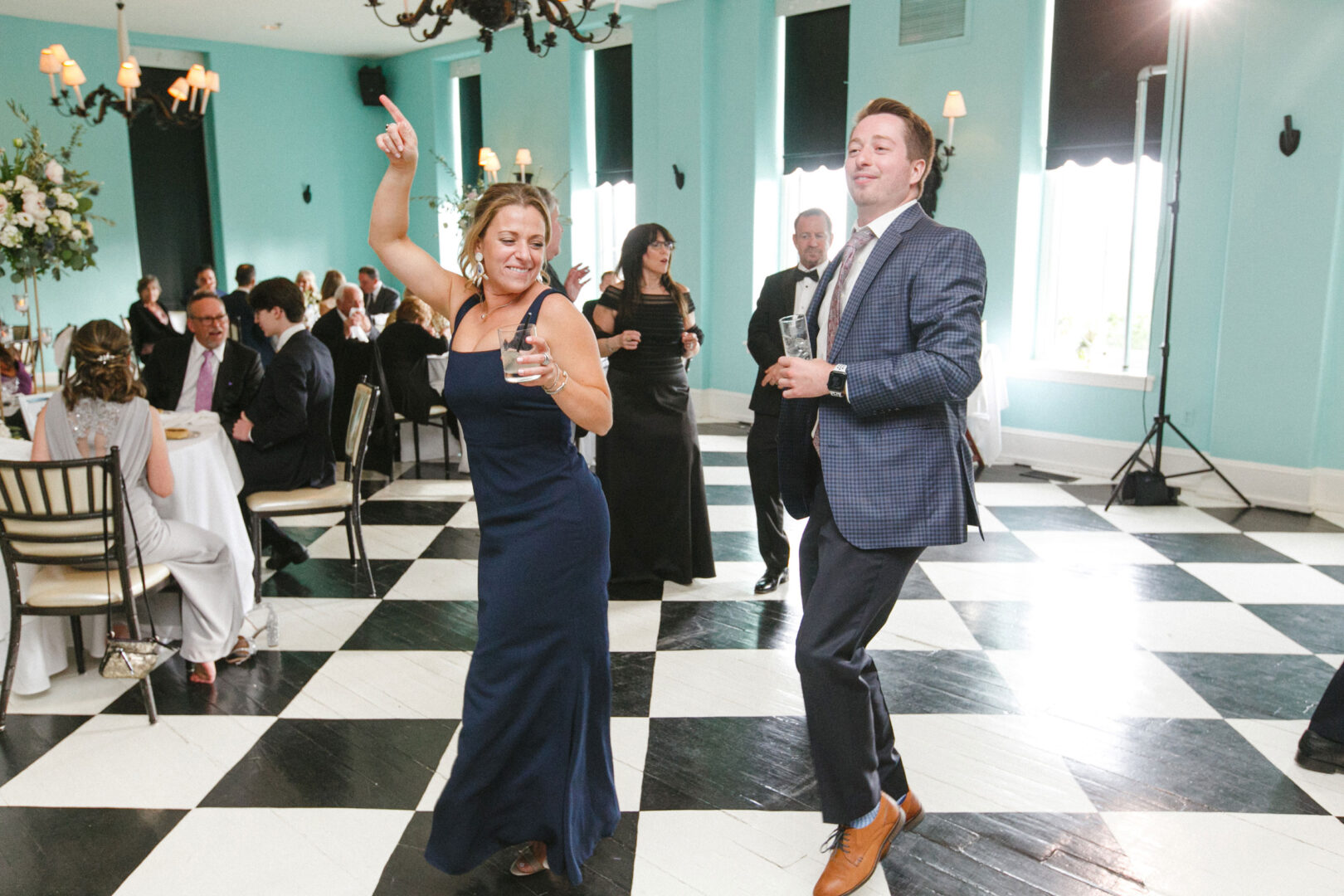 At a wedding in Cape May at Congress Hall, two people in formal attire dance energetically on a black and white checkered floor, their joyous movements captivating the seated guests in the background.