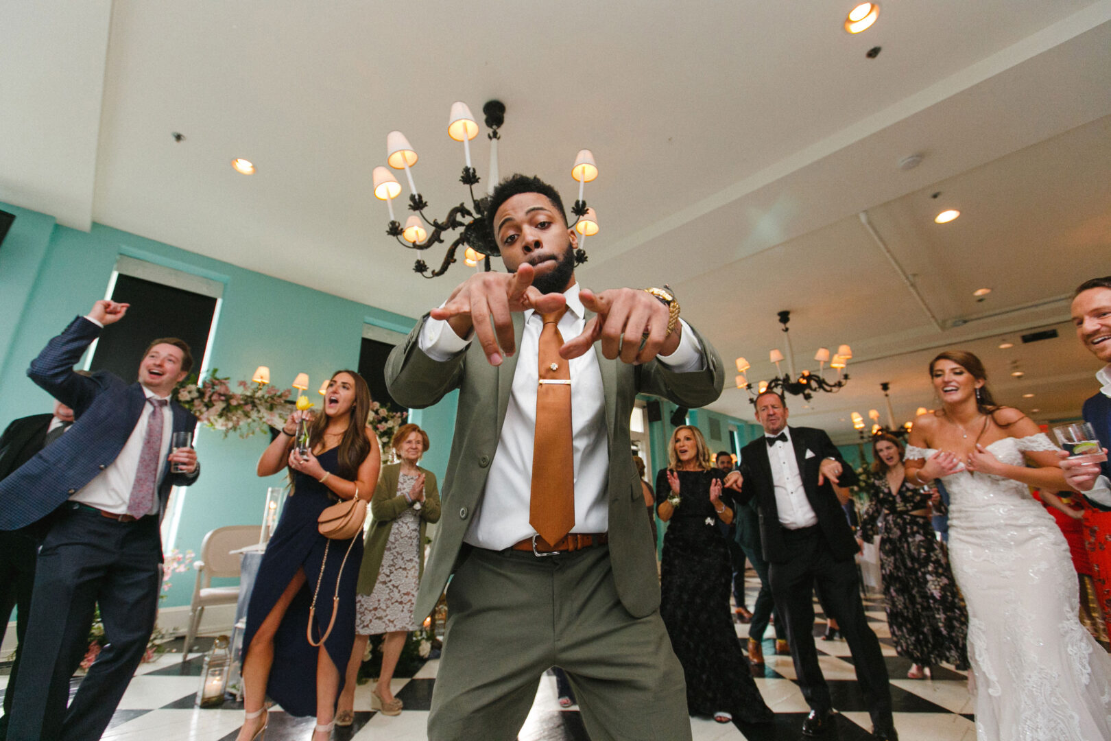 At a wedding in Cape May at Congress Hall, a group of people dances energetically indoors. One person in the foreground points playfully to the camera, surrounded by a room featuring a chandelier and black-and-white checkered floor.