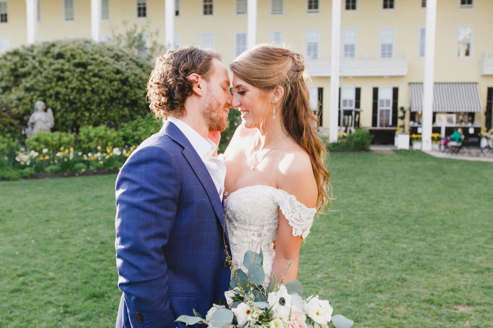 A couple in formal attire embraces joyfully, celebrating their wedding in Cape May at the iconic Congress Hall, with its large yellow facade complemented by green lawns and shrubs.