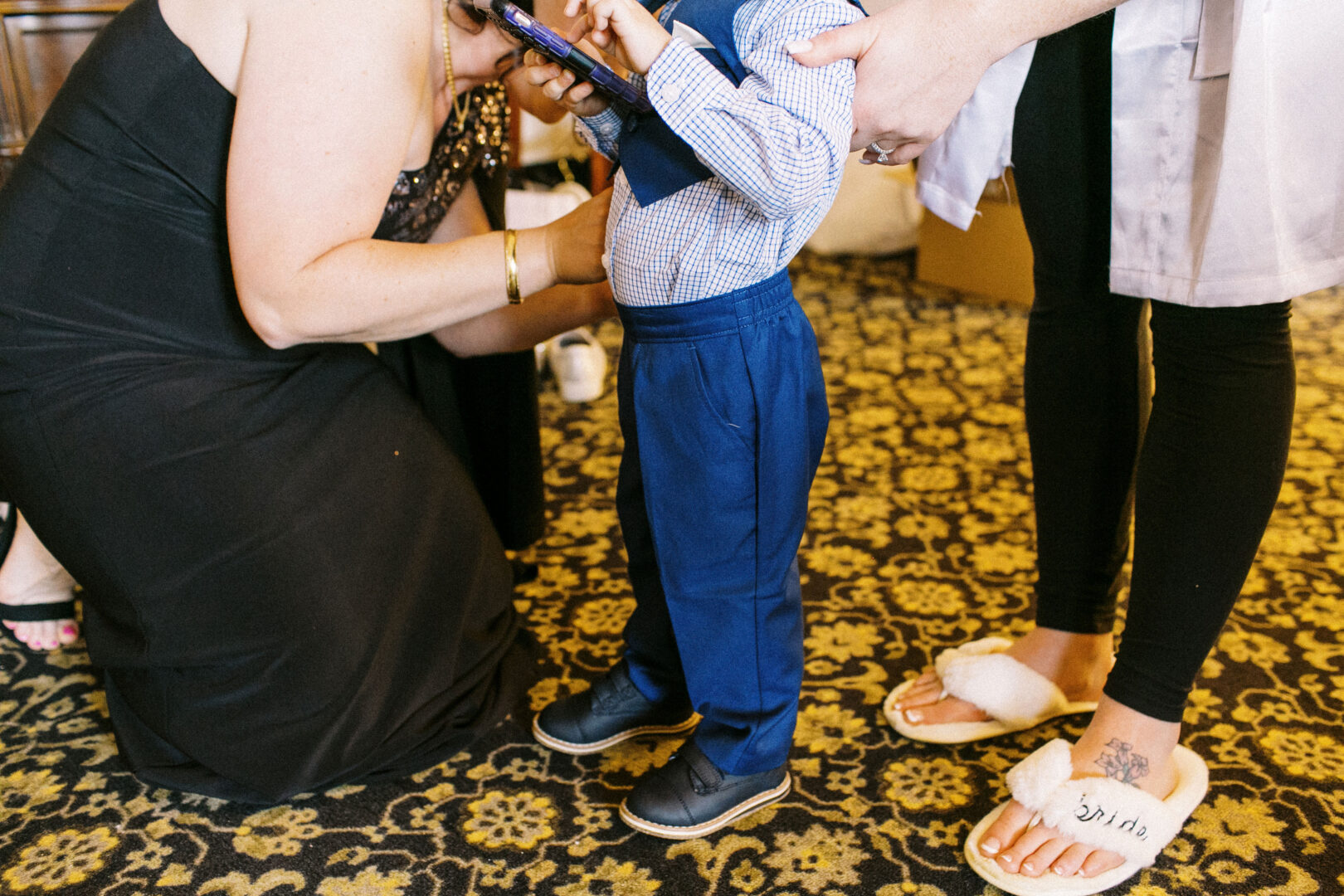 At an Atlantic City wedding, a child in formal attire is being assisted by an adult in a black dress, while another person in slippers stands nearby on the patterned carpet.