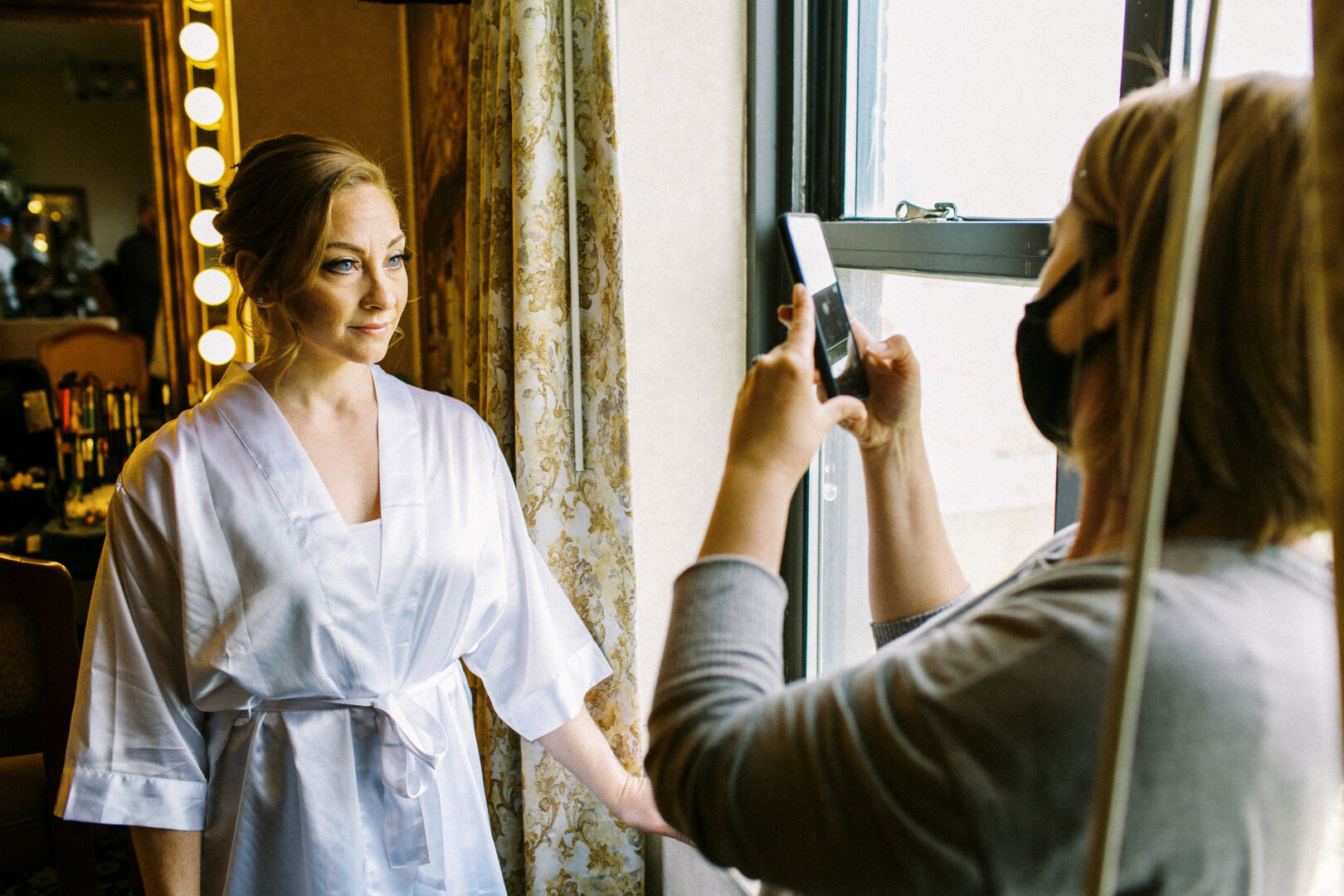 A woman in a white robe poses near a window, capturing the essence of an Atlantic City wedding, while another person takes her photo with a smartphone inside the room.