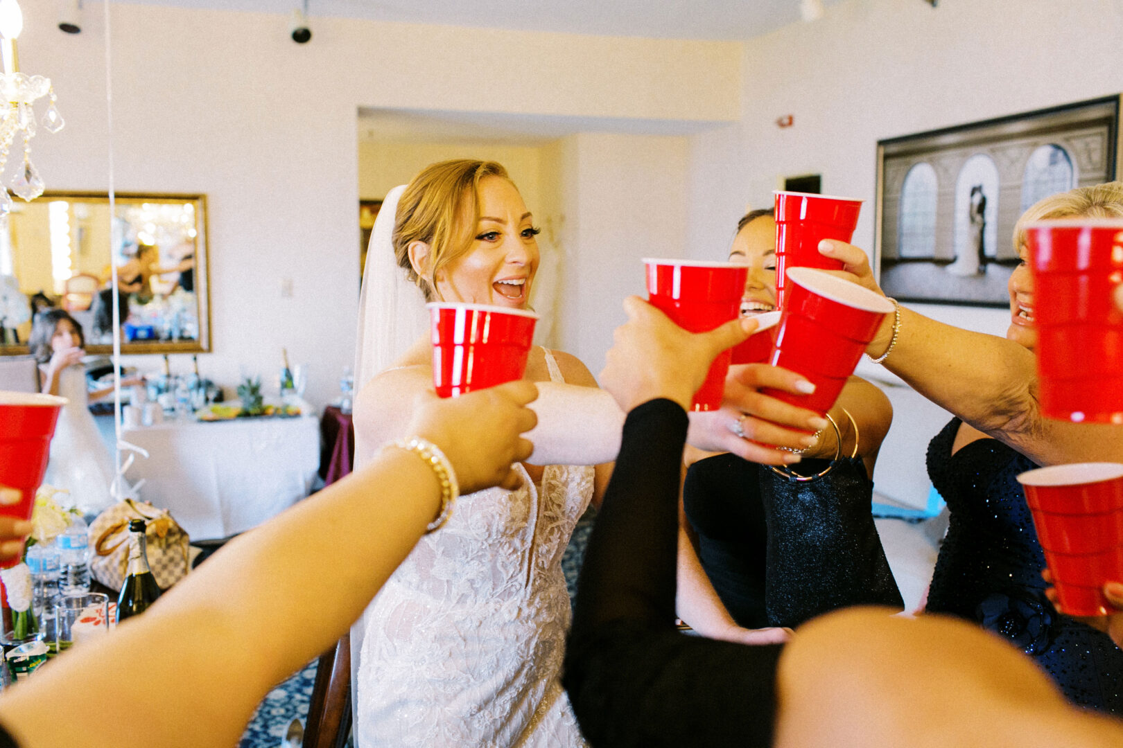 A bride in a white dress toasts with friends holding red cups in a beautifully decorated room, capturing the joyous spirit of an Atlantic City wedding.
