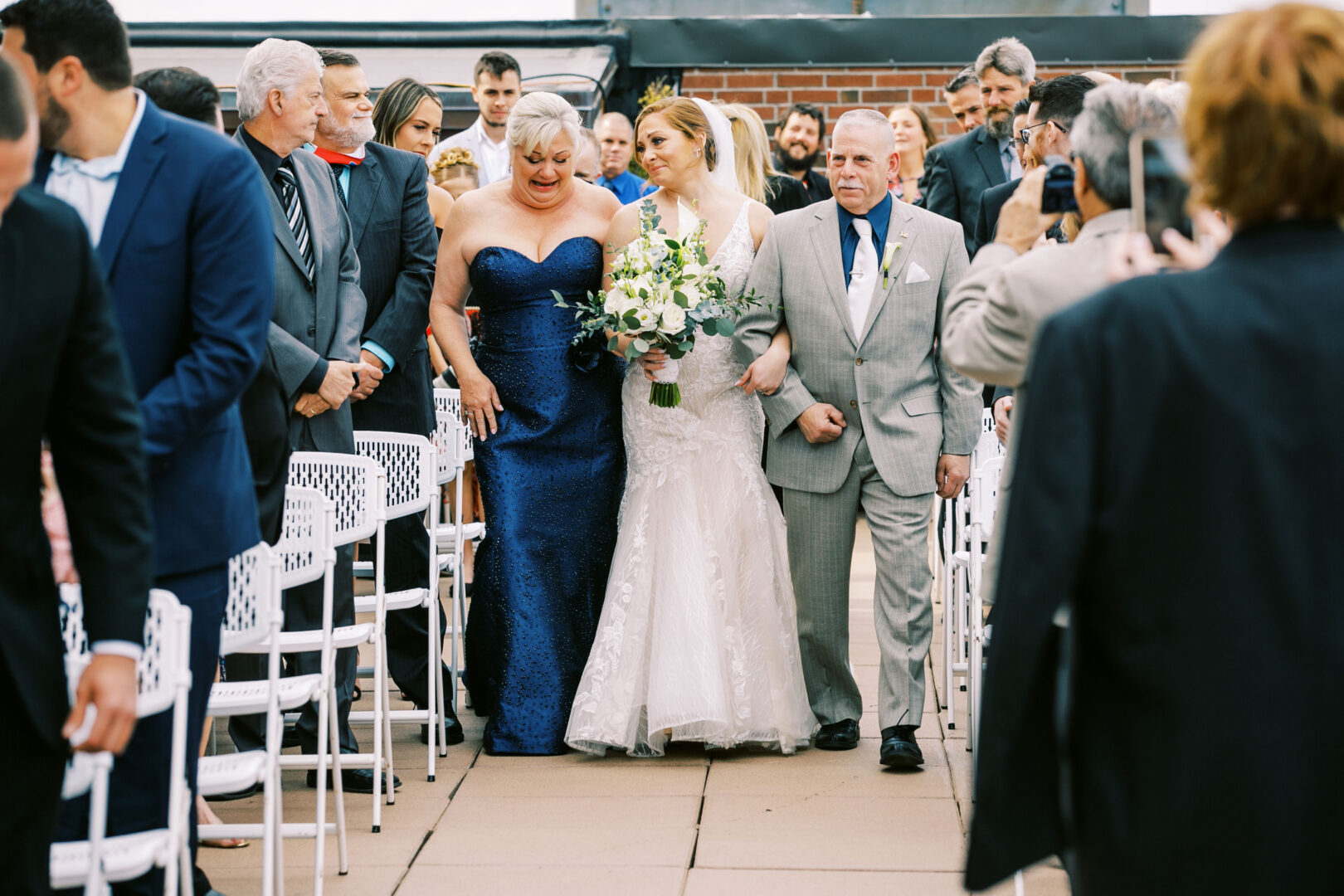 The bride, in a stunning white dress, gracefully walks down the outdoor aisle with two older adults at her side, surrounded by seated and standing guests. The serene backdrop of an Atlantic City wedding adds a touch of enchantment to this unforgettable moment.