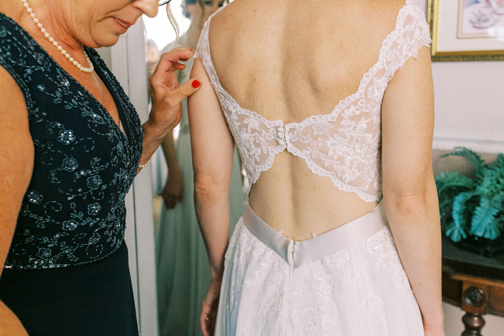 A woman in a lace-backed dress has her attire adjusted by another person in a dark, embellished gown. A mirror reflects this elegant scene, capturing the essence of an Old York Road Country Club wedding.