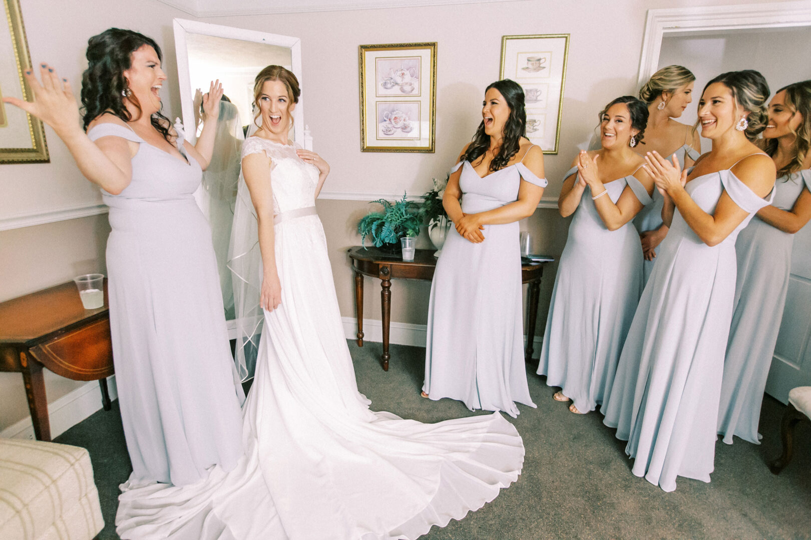 At the Old York Road Country Club wedding, a bride in a white dress poses gracefully while bridesmaids in matching light blue dresses applaud and smile, framed by elegant artwork and a lush potted plant.