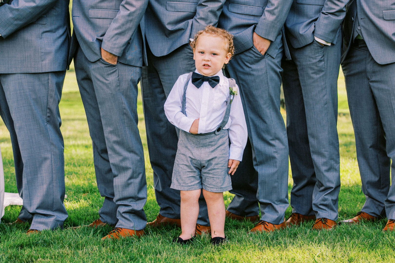 A small child, dressed in a bow tie, suspenders, and shorts, stands adorably on the lush grass at an Old York Road Country Club wedding, in front of a row of adults in gray suits and brown shoes.