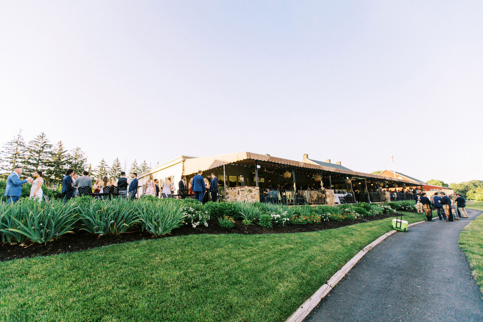 Guests mingling outside a large building at the Old York Road Country Club wedding, with a tented area surrounded by lush green lawns and gardens under a clear sky.