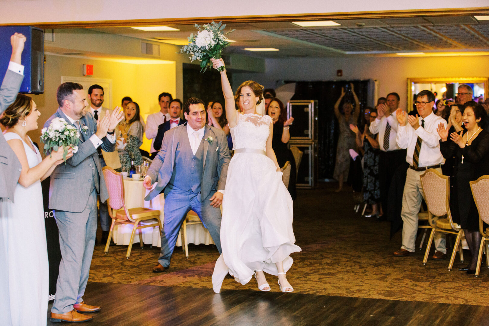 A bride and groom enter the Old York Road Country Club enthusiastically, holding hands. The bride raises her bouquet in the air. Guests around them cheer and clap.