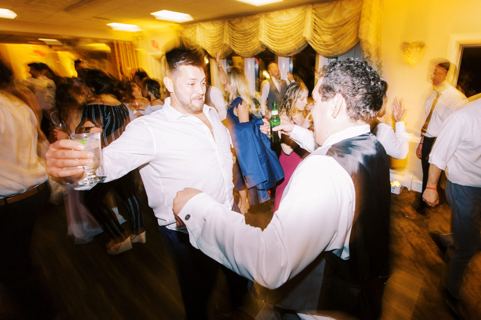 Two men in white shirts are joyfully dancing and holding drinks, celebrating in a lively indoor party setting at an Old York Road Country Club wedding.