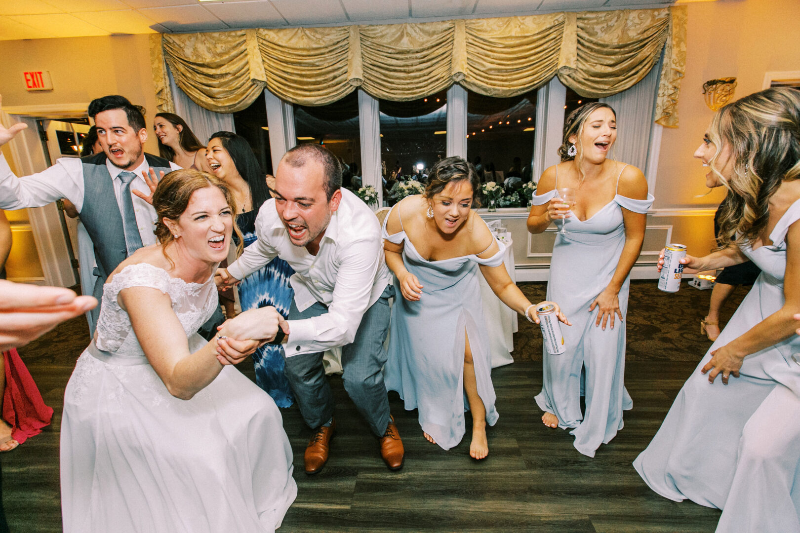At the Old York Road Country Club wedding, the party joyfully dances indoors, with people laughing and holding drinks. The bride and groom are at the center, surrounded by bridesmaids in light blue dresses.