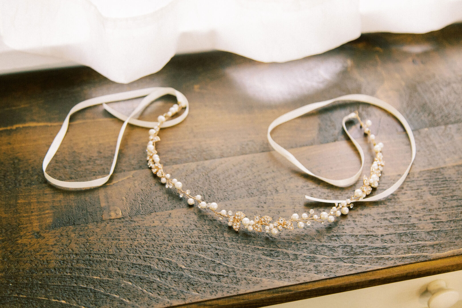 A delicate beaded headband with pearls and gold accents lies on a wooden surface, reminiscent of the rustic elegance of a wedding at Harvest View Barn at Hershey Farms, with long cream ribbons gracefully attached.