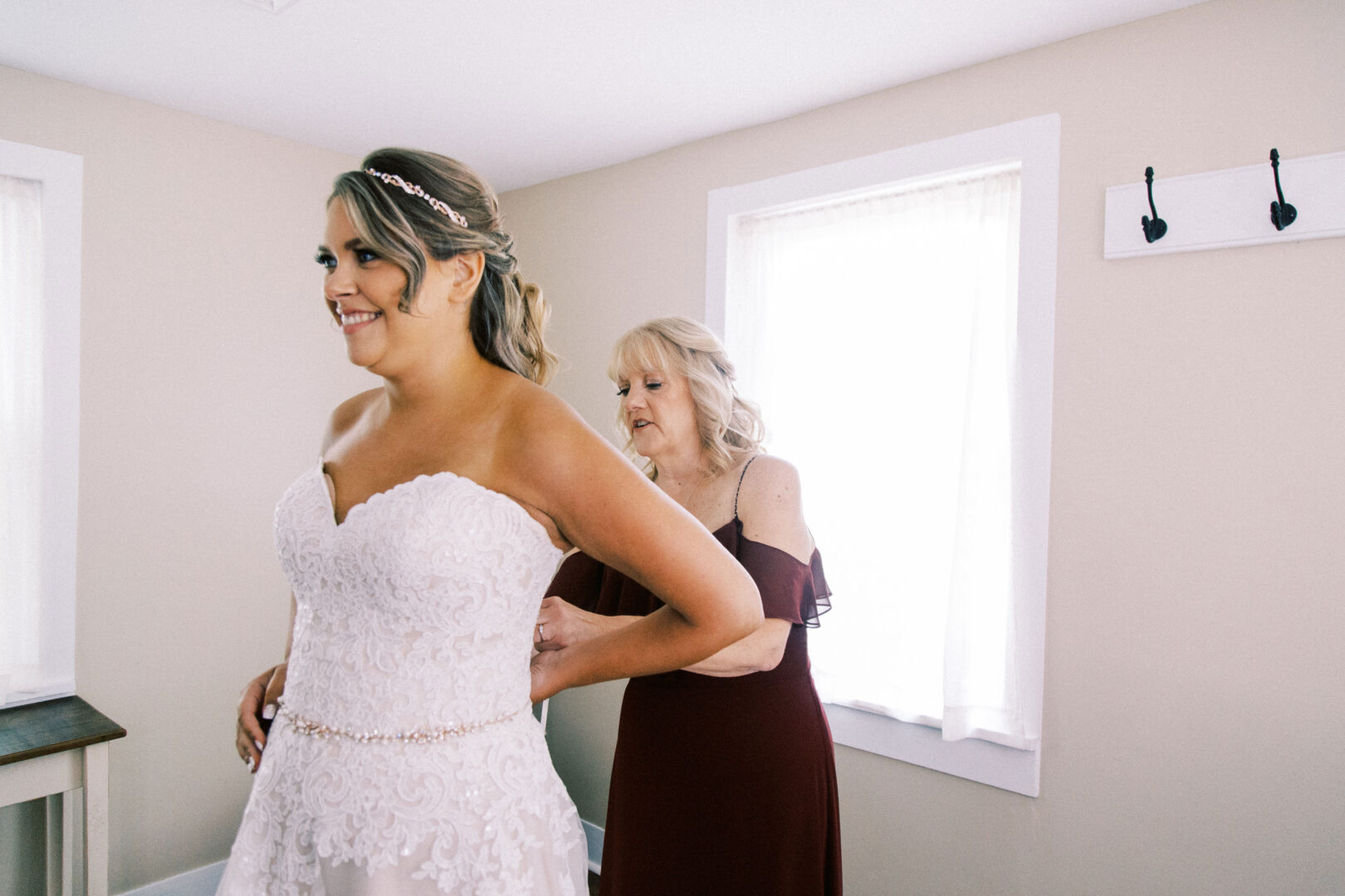 In a bright room at Harvest View Barn, a woman assists the bride in adjusting her strapless lace wedding dress.