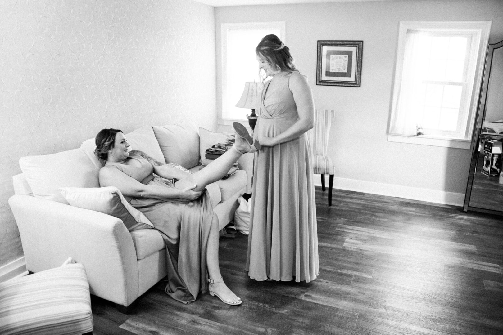 In the cozy living room, one woman lounges on the couch with her legs up, while the other gently holds her foot. The bright space, reminiscent of a Harvest View Barn at Hershey Farms wedding, features a warm lamp and an elegant framed picture on the wall.