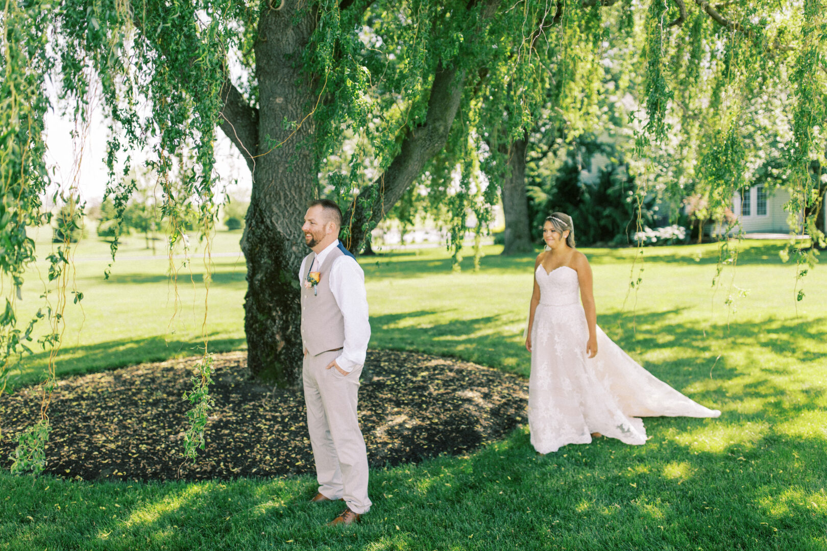 The bride gracefully approaches the groom standing under a large tree in formal attire at their enchanting Harvest View Barn at Hershey Farms wedding, both surrounded by lush greenery.