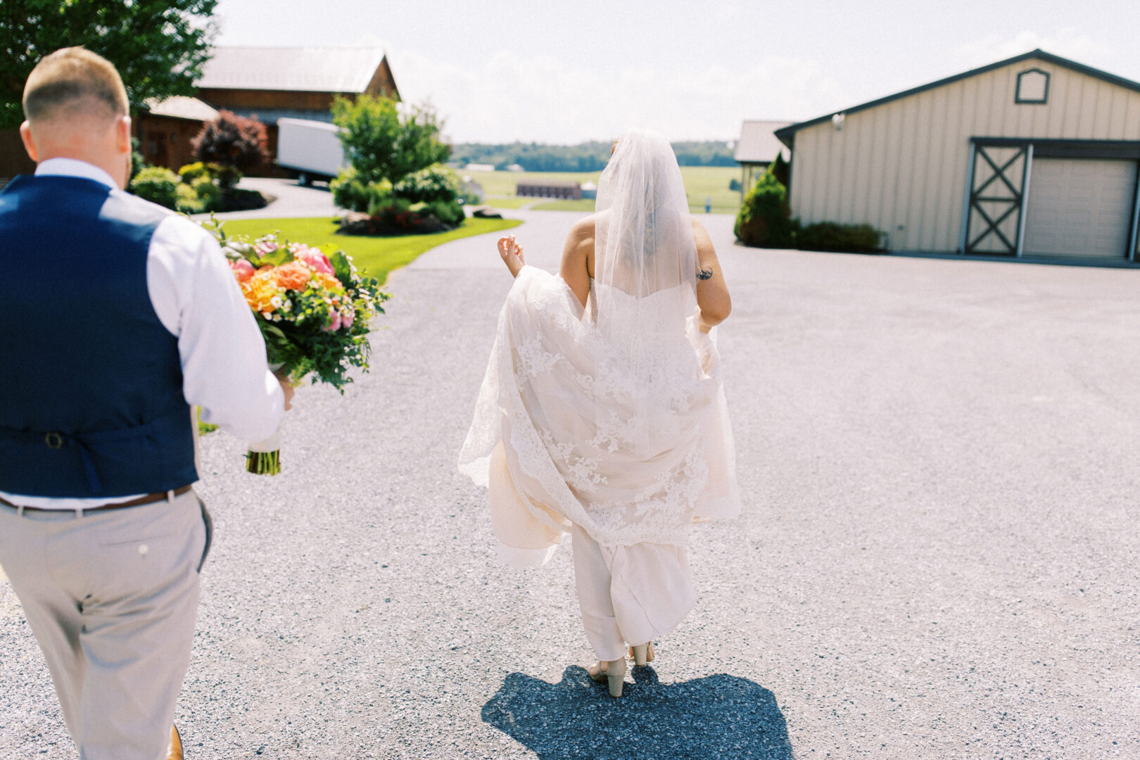 A bride in a wedding dress walks along the gravel path, holding her gown, as the groom in a vest follows with a bouquet. The lush greenery and barns of Harvest View Barn at Hershey Farms set the perfect backdrop for this enchanting wedding scene.