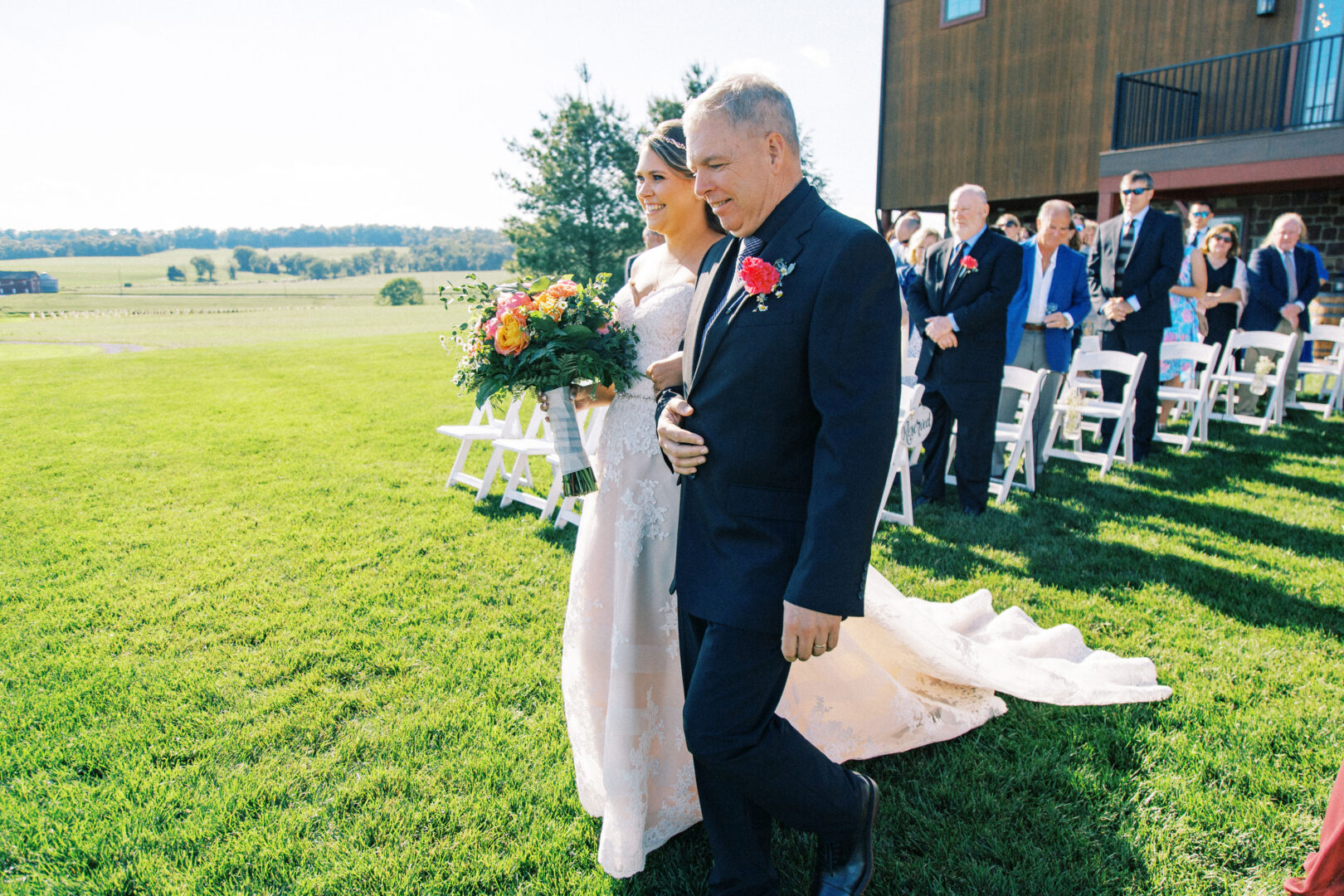 At the Harvest View Barn at Hershey Farms wedding, a bride in a white dress walks down an outdoor aisle with an older man in a suit, holding a bouquet of flowers. Guests are seated on white chairs under the clear sky, witnessing this beautiful moment.