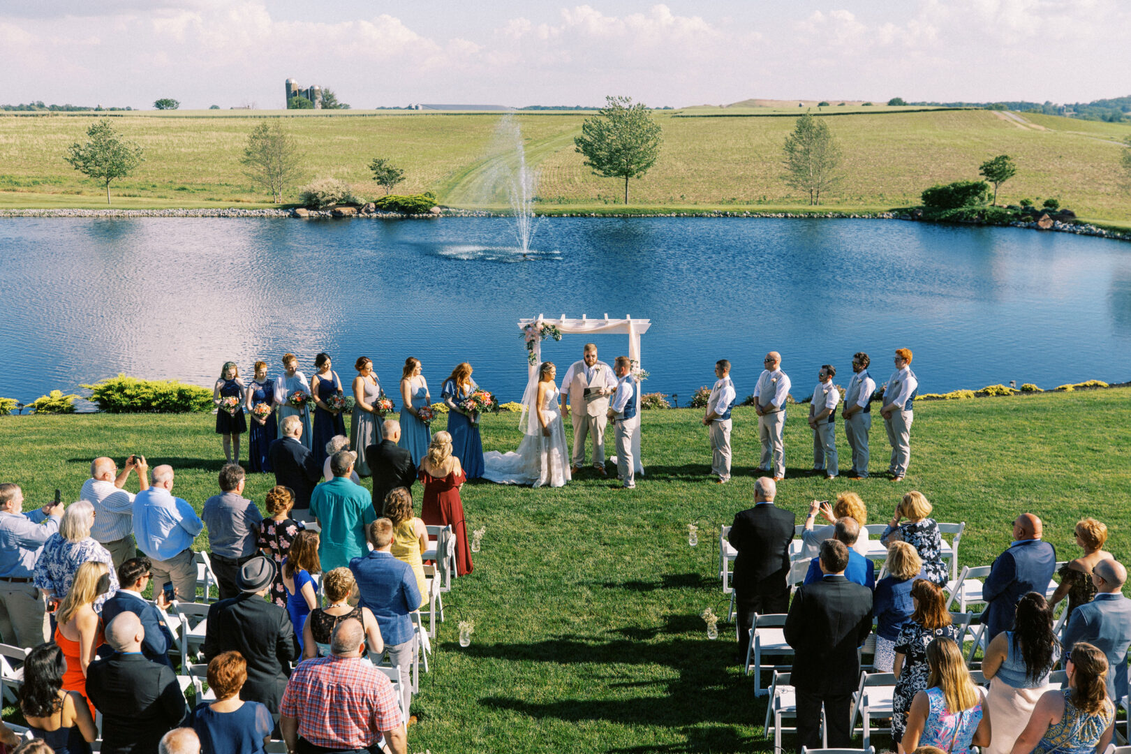 A wedding ceremony outdoors by a lake at Harvest View Barn at Hershey Farms. Guests are seated, and the wedding party stands under an arbor.