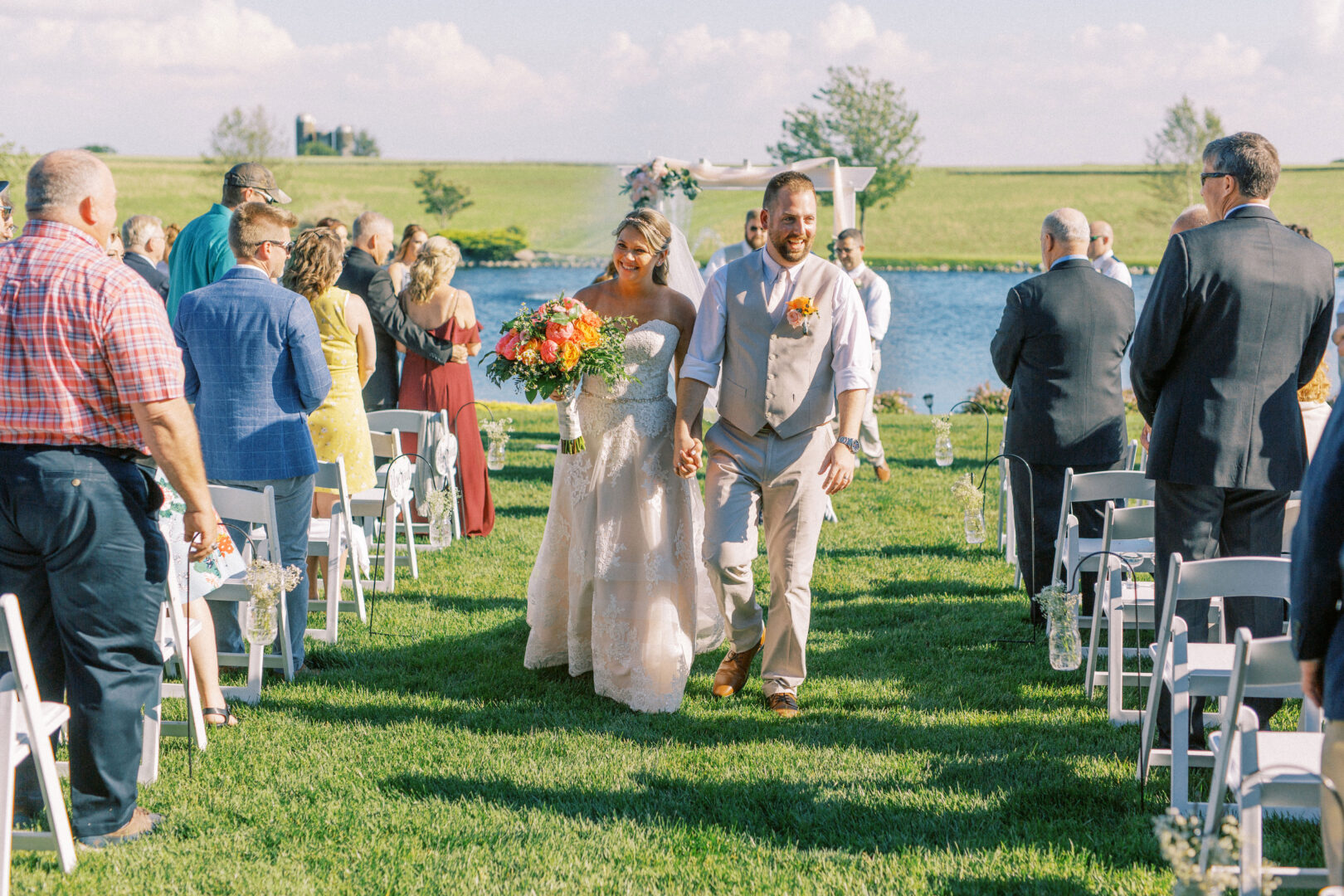 At Harvest View Barn at Hershey Farms, a bride and groom walk down an outdoor aisle, smiling and holding hands, as guests seated on white chairs watch. A serene lake and lush trees create the perfect backdrop for this enchanting wedding moment.