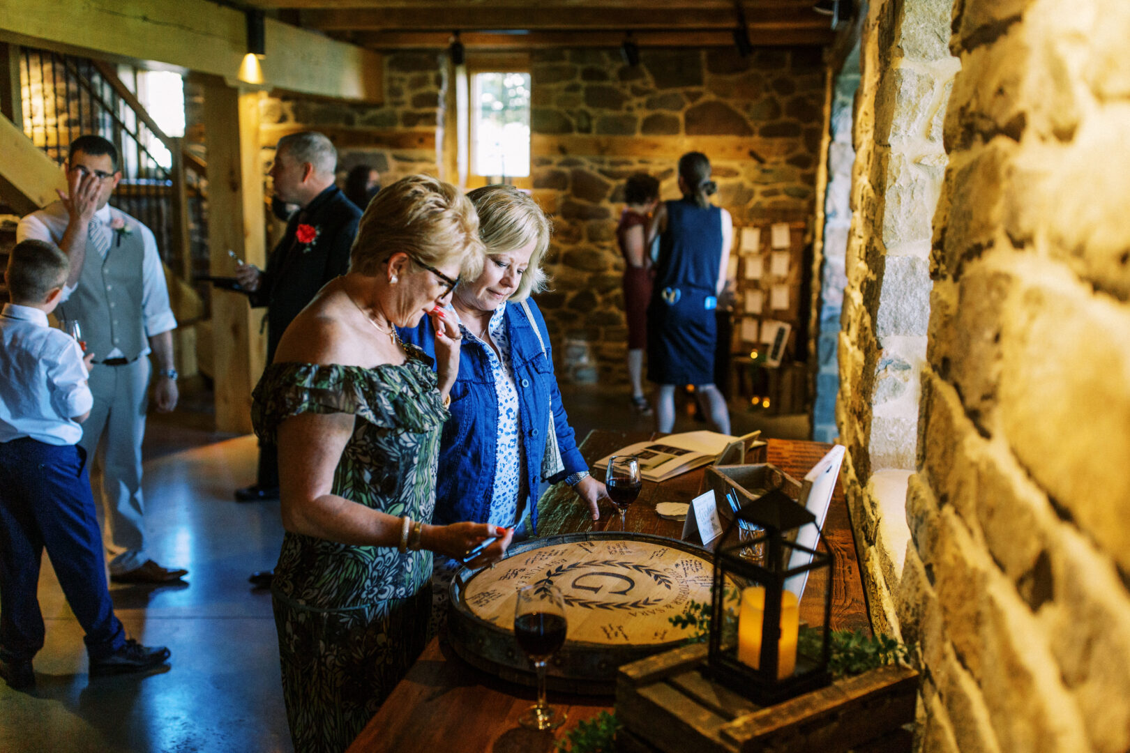 Two women stand at a wooden table in Harvest View Barn at Hershey Farms, examining a circular item during a social gathering in the rustic room with stone walls. Guests are mingling in the background, creating a lively atmosphere at this charming countryside venue.