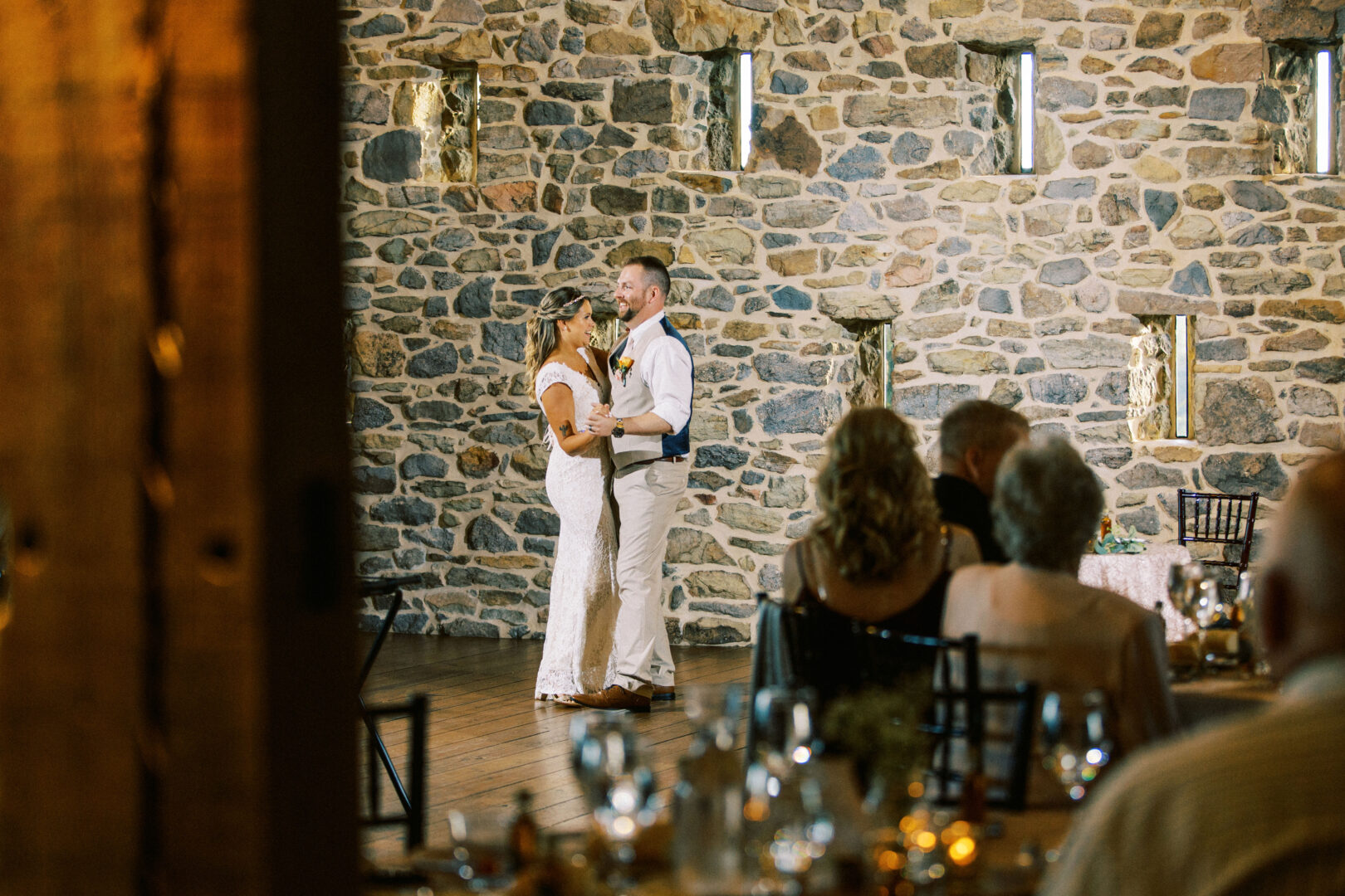 At the Harvest View Barn at Hershey Farms wedding, a couple dances together at the reception, surrounded by guests seated at tables. The charming stone wall in the background adds to the enchanting atmosphere.