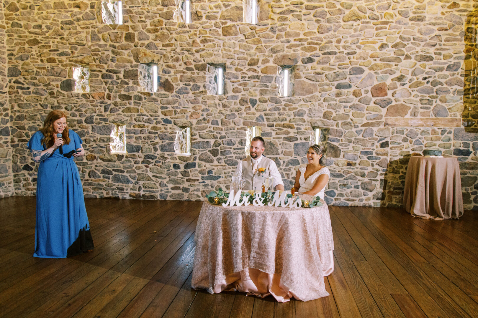 A couple sits at a table with a "Mr & Mrs" sign, basking in the cozy ambiance of the Harvest View Barn at Hershey Farms. A woman in a blue dress stands nearby, passionately giving a speech against the charming stone wall background.