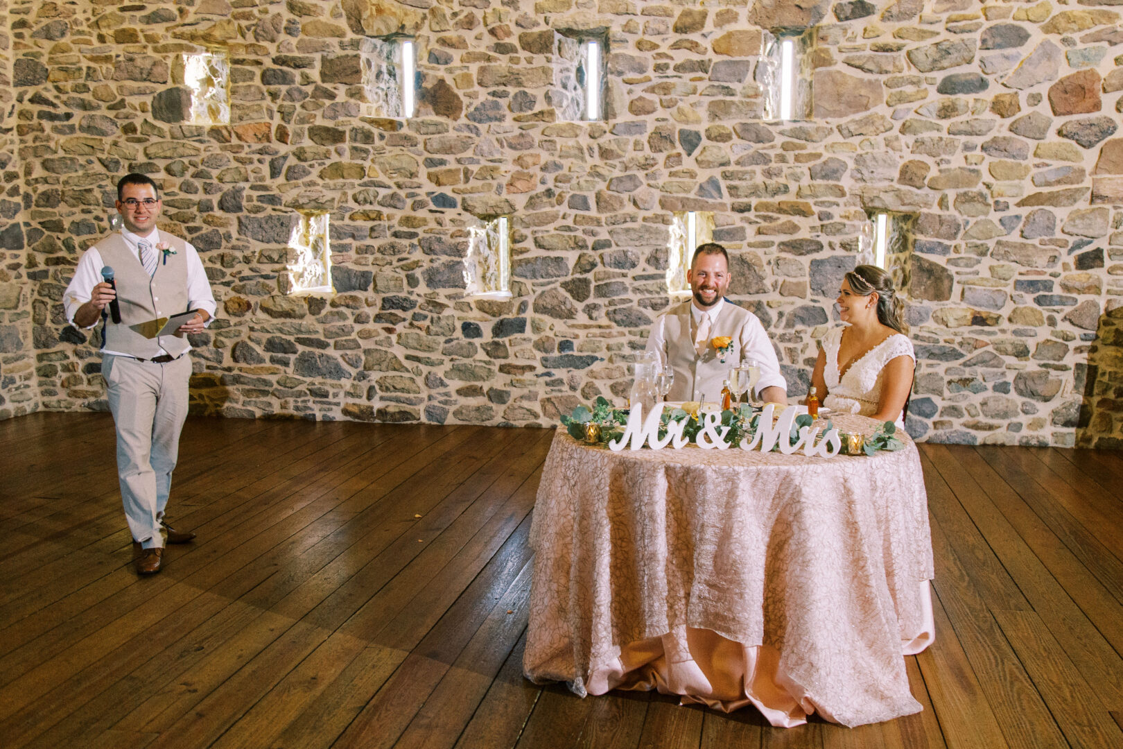 A couple sits at a round table labeled "Mr. & Mrs." in front of a stone wall at Harvest View Barn, while a person stands nearby holding a notebook.