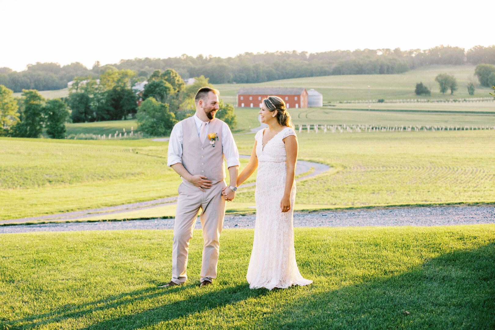 A couple in formal attire stands on a grassy field at Harvest View Barn at Hershey Farms Wedding, holding hands against a rural backdrop featuring a charming red barn and lush trees.