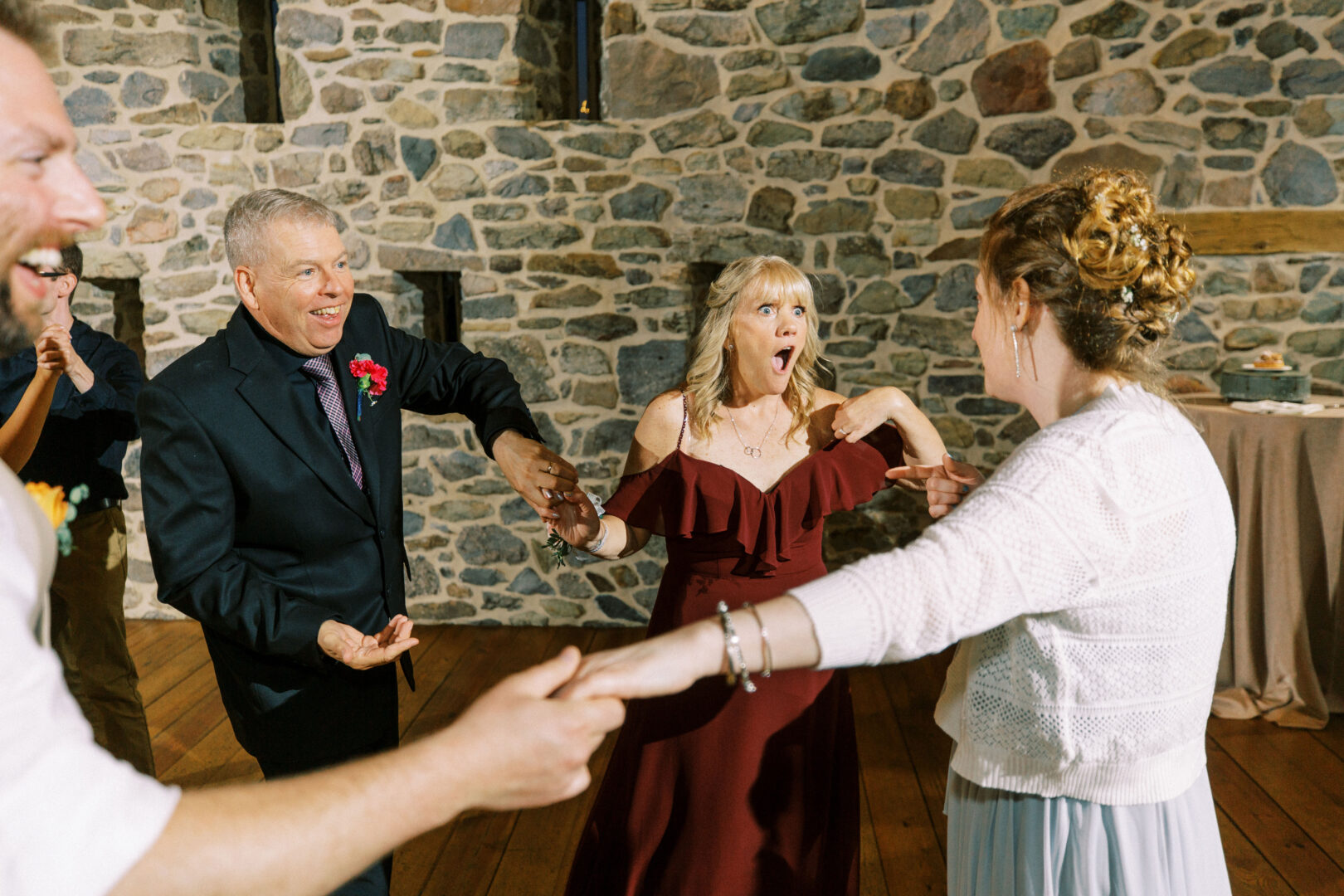 A group of people holding hands in a circle, with one woman in a red dress looking surprised, gathers joyfully at Harvest View Barn at Hershey Farms Wedding. They are set against a charming stone wall backdrop, adding rustic elegance to the joyful moment.